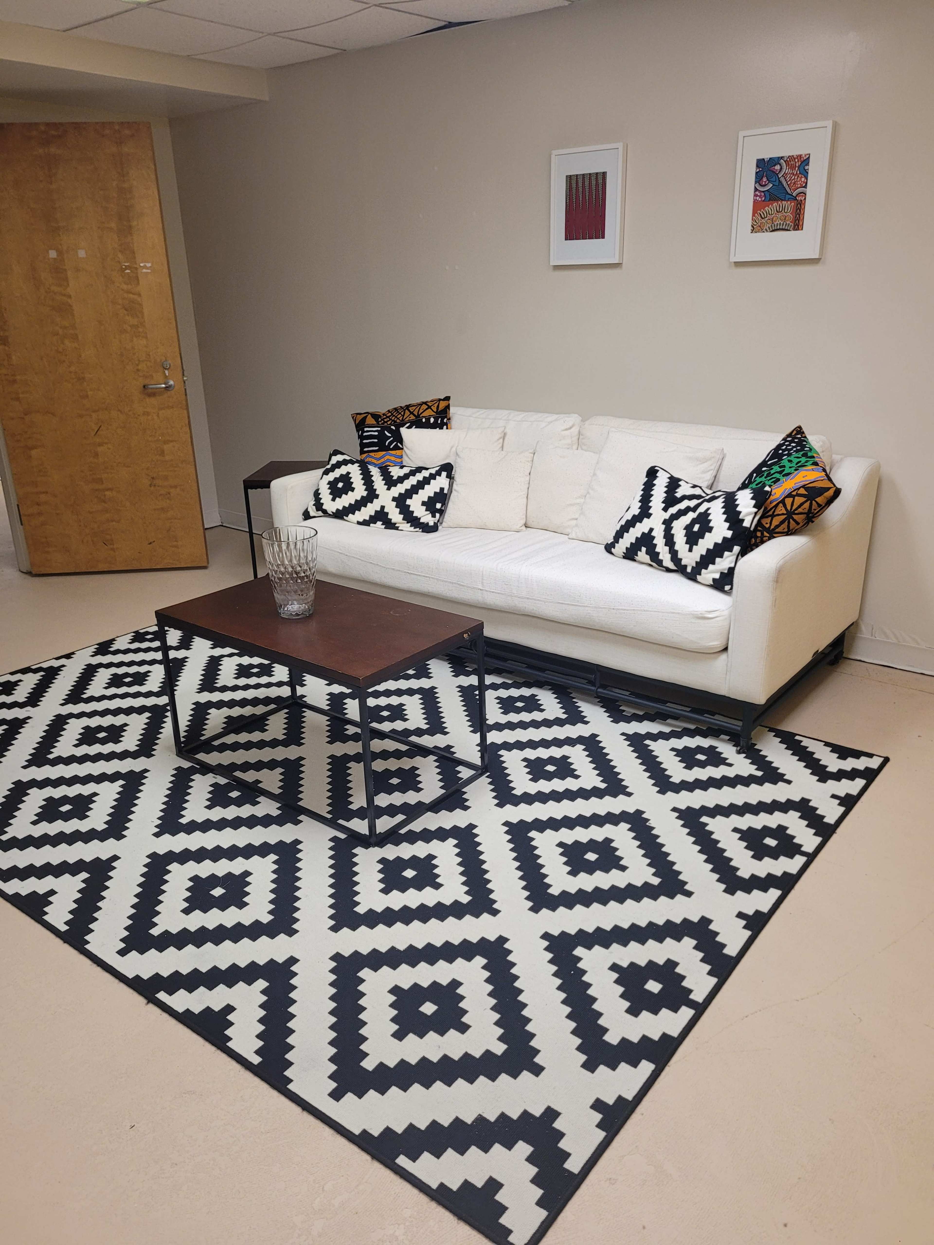 The image shows a living room area featuring a white sofa with patterned cushions, a wooden coffee table, and a geometric-patterned rug on the floor.