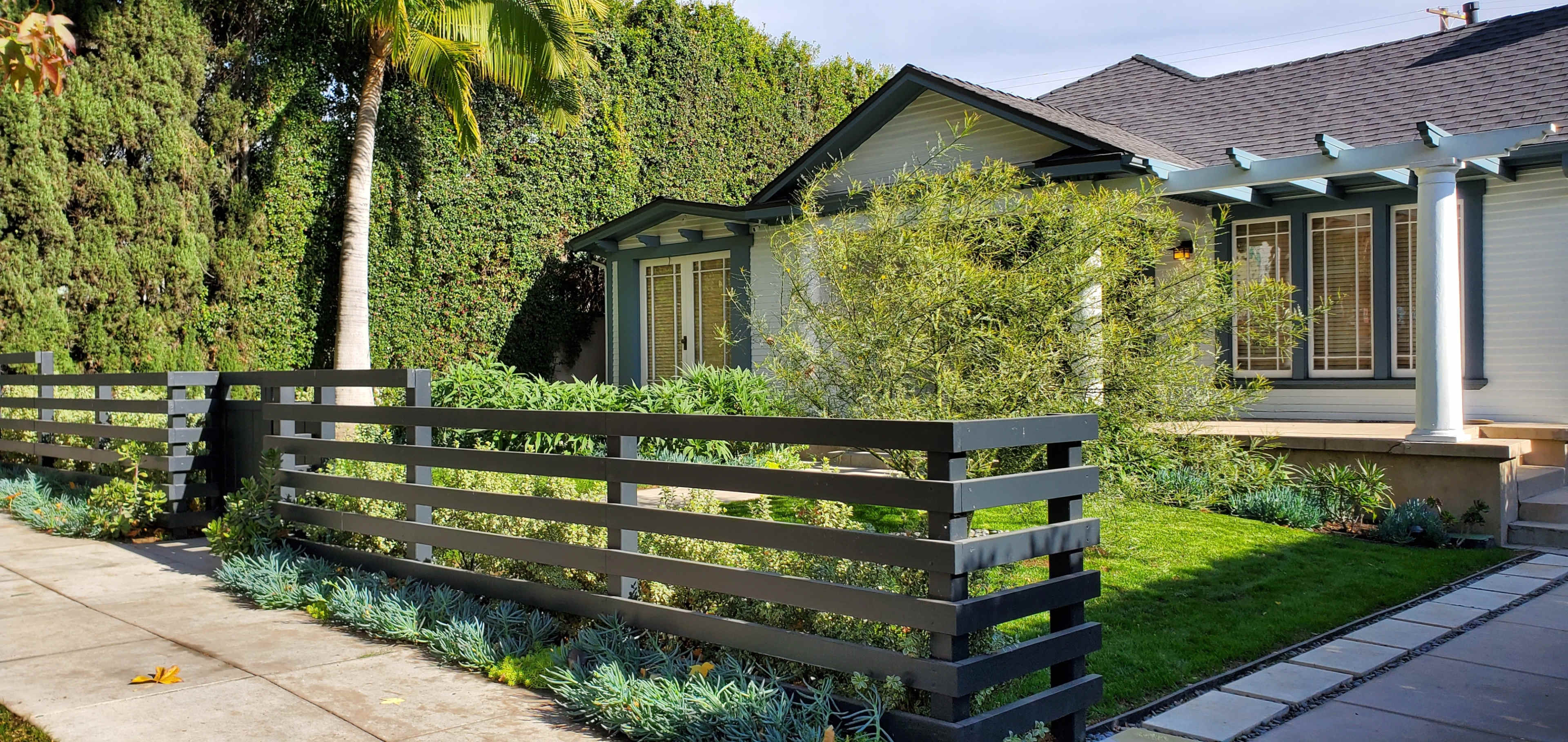 A landscaped front yard with a modern fence and a light-colored house surrounded by trees and shrubs.