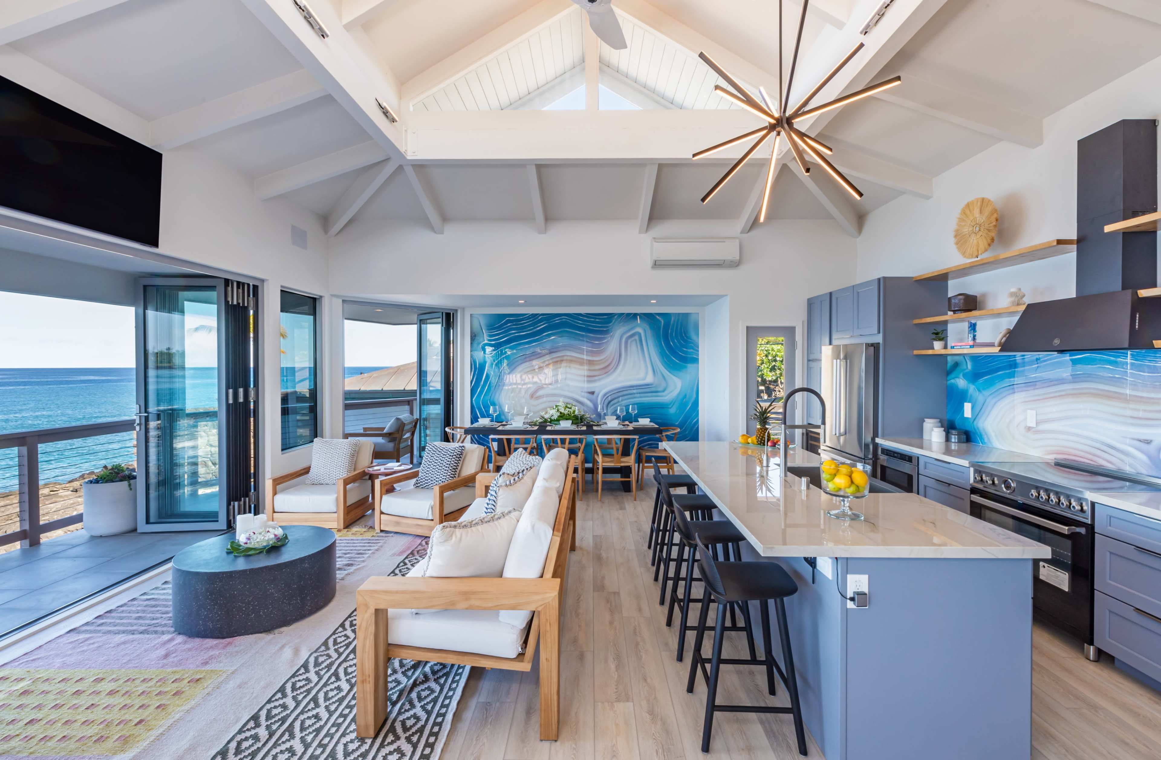 A modern open-concept kitchen and living area with large windows overlooking the ocean, featuring light-colored furnishings and a striking blue wall art.
