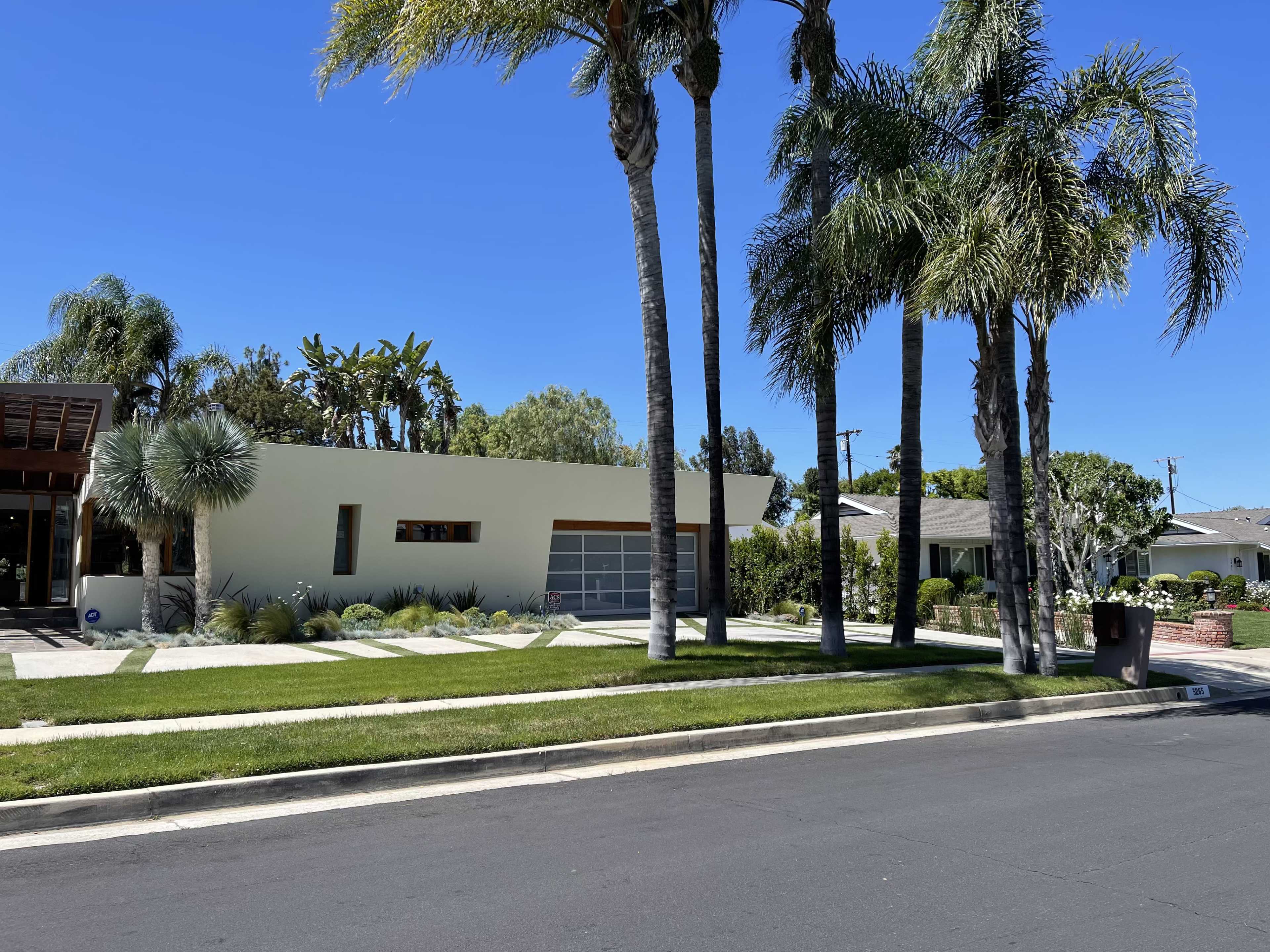 A modern house with a garage, surrounded by palm trees and well-maintained landscaping, on a residential street.