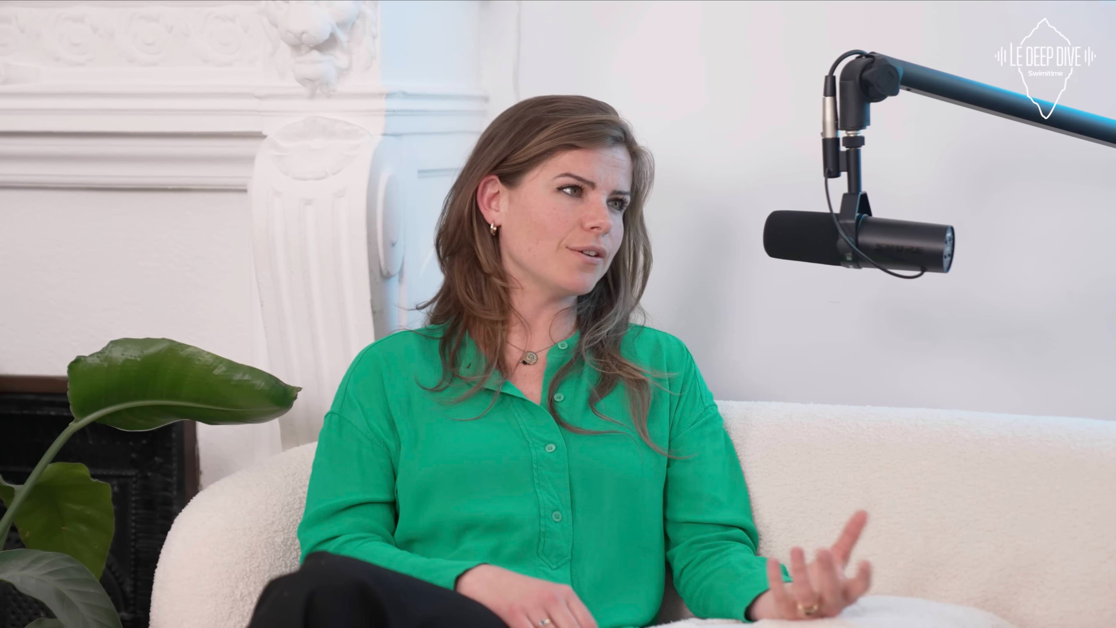 A woman with long hair is sitting on a couch, speaking into a microphone in a well-lit room with a plant nearby.