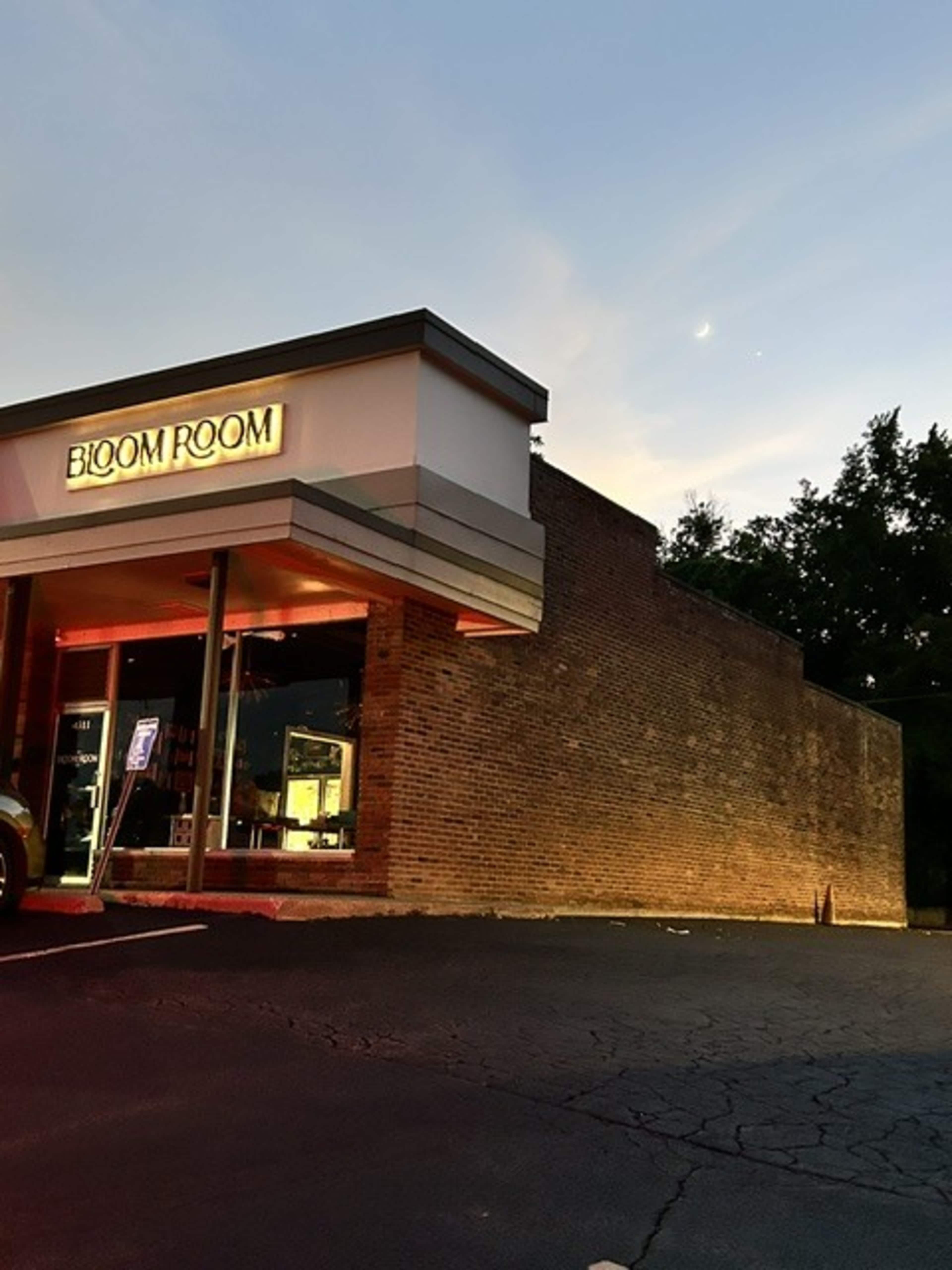 The image shows a building with the sign "BLOOM ROOM" illuminated at dusk, alongside a brick wall and a parking area.