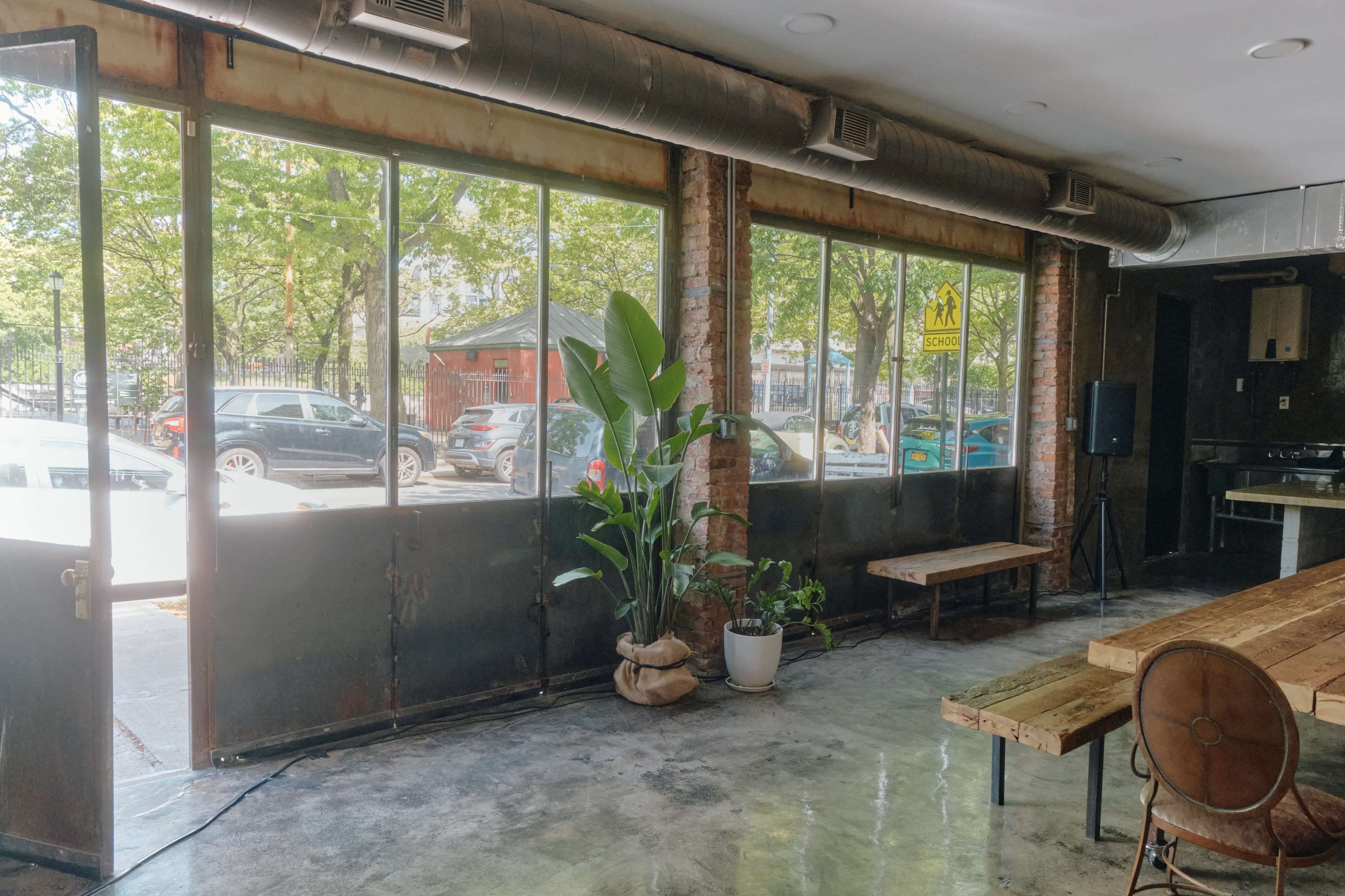 The interior of a spacious café features large windows, brick walls, a wooden table, and a potted plant near the entrance.