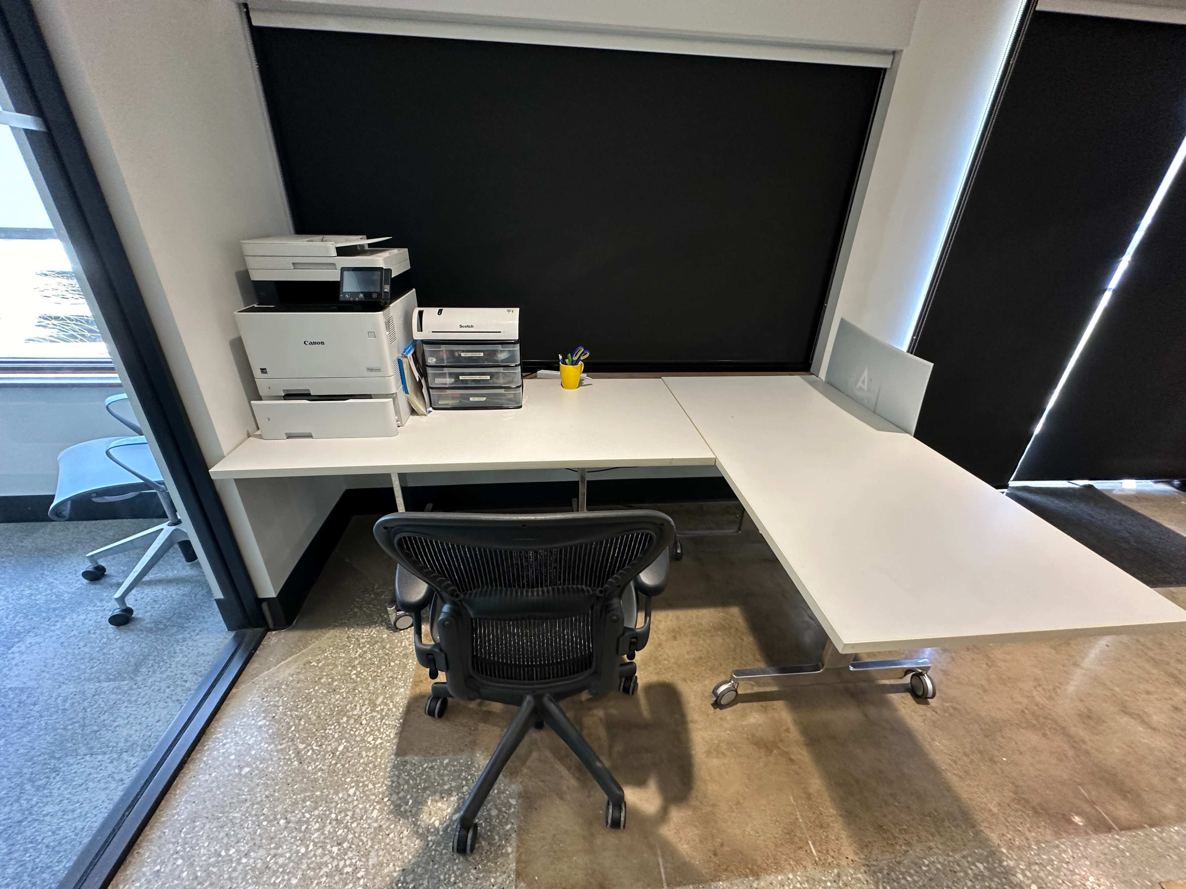 A clean office workspace with a black rolling chair, a white desk, a printer, and a yellow container holding pens.