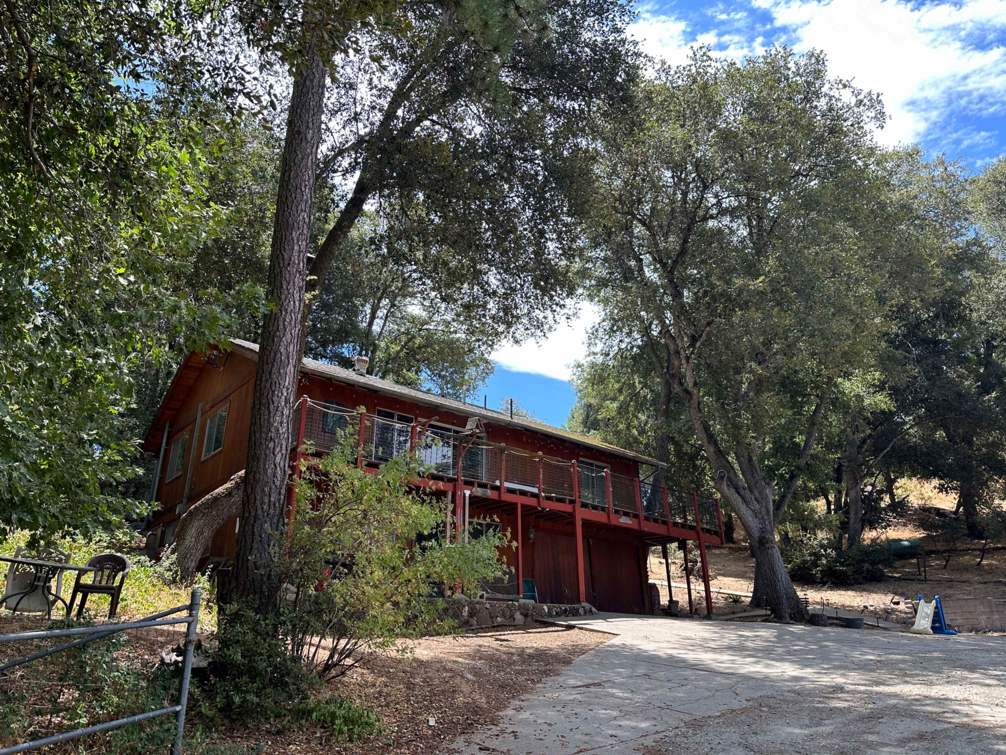 A two-story red wooden house with a balcony overlooks a gravel driveway surrounded by trees and hilly terrain.