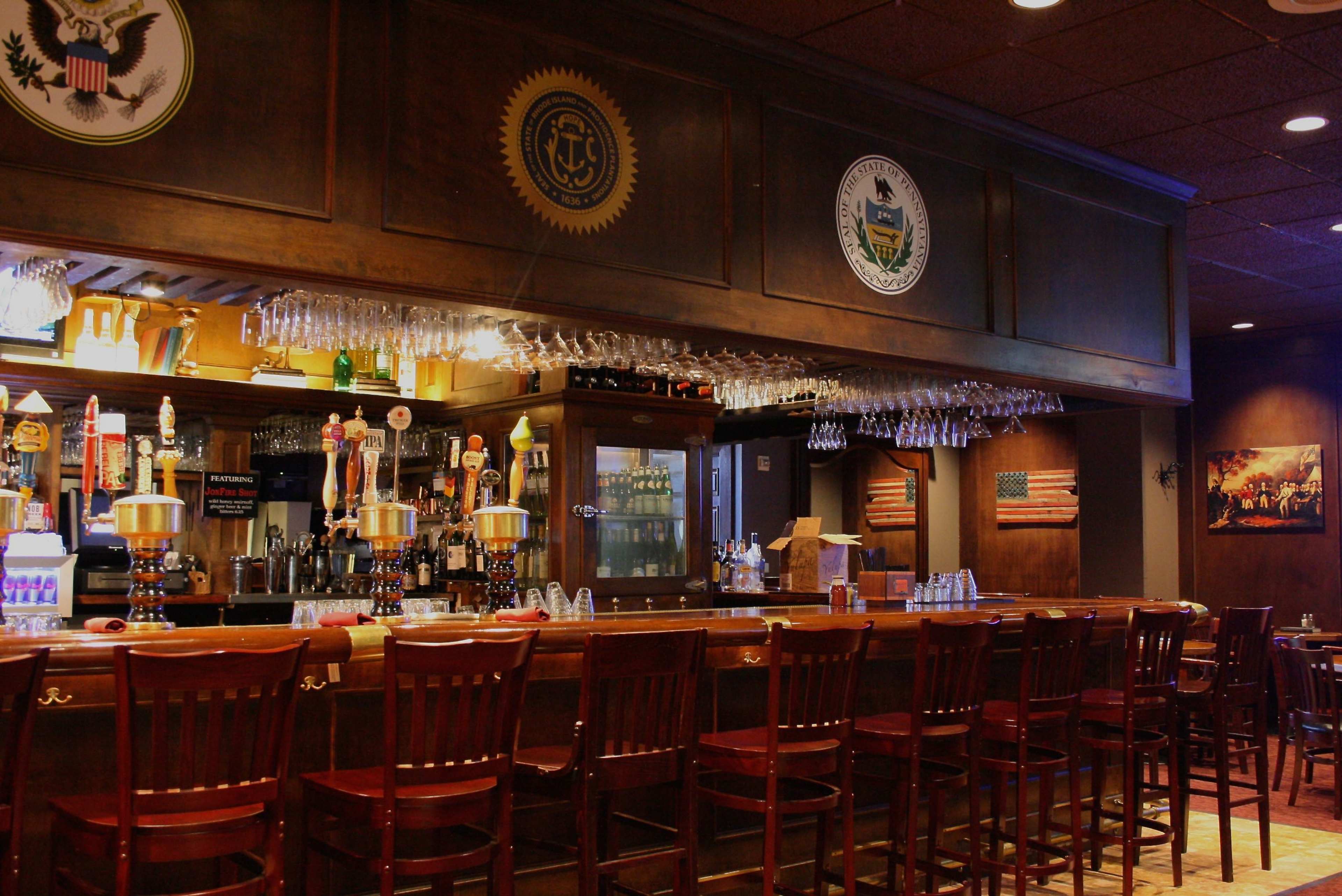 A wooden bar with several beer taps and a row of high-backed chairs, illuminated by soft lighting and decorated with emblems and flags on the walls.