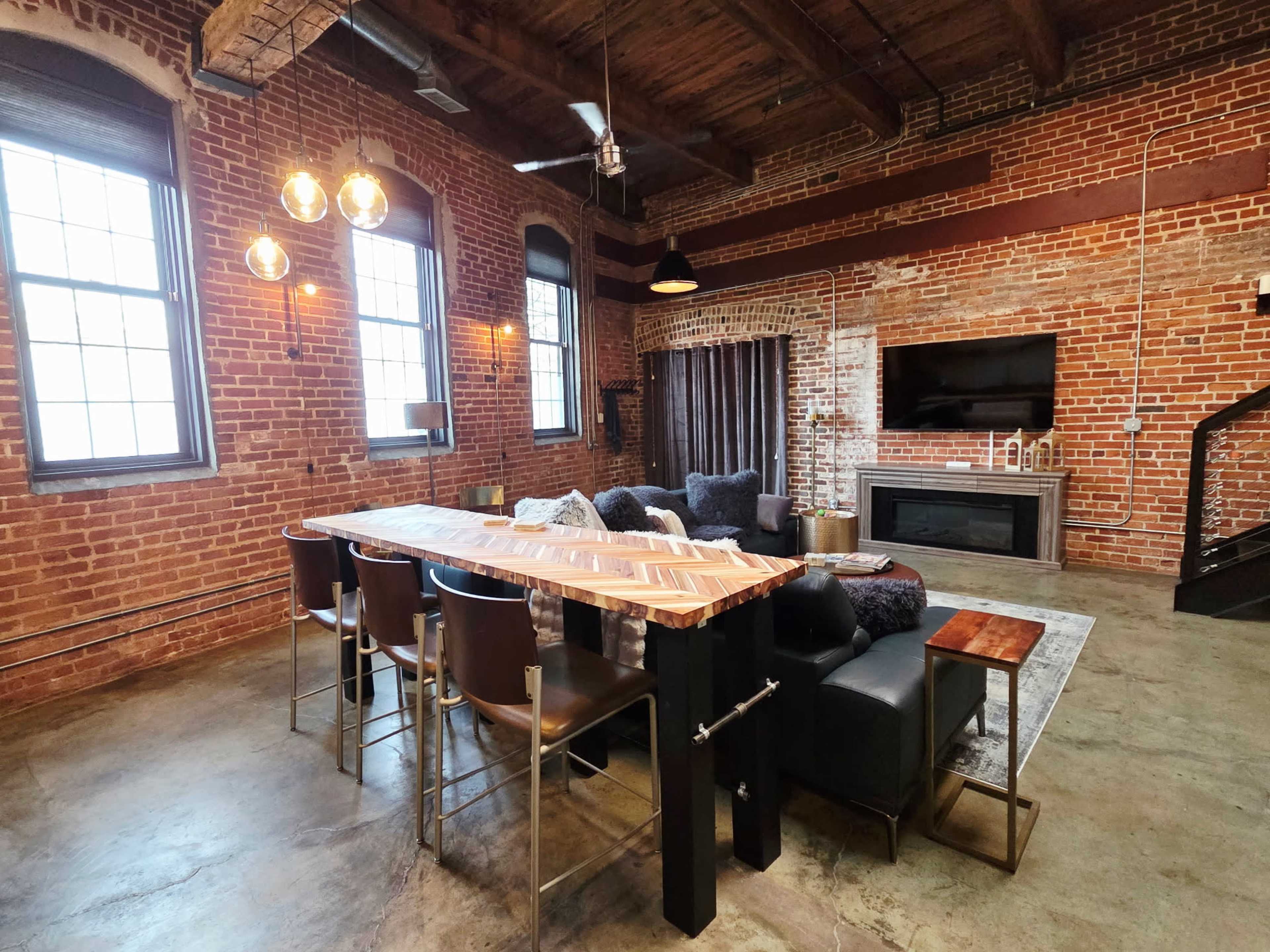The image shows an industrial-style loft with exposed brick walls, large windows, a dining area with a wooden table, and a cozy living space featuring a fireplace and a modern entertainment setup.