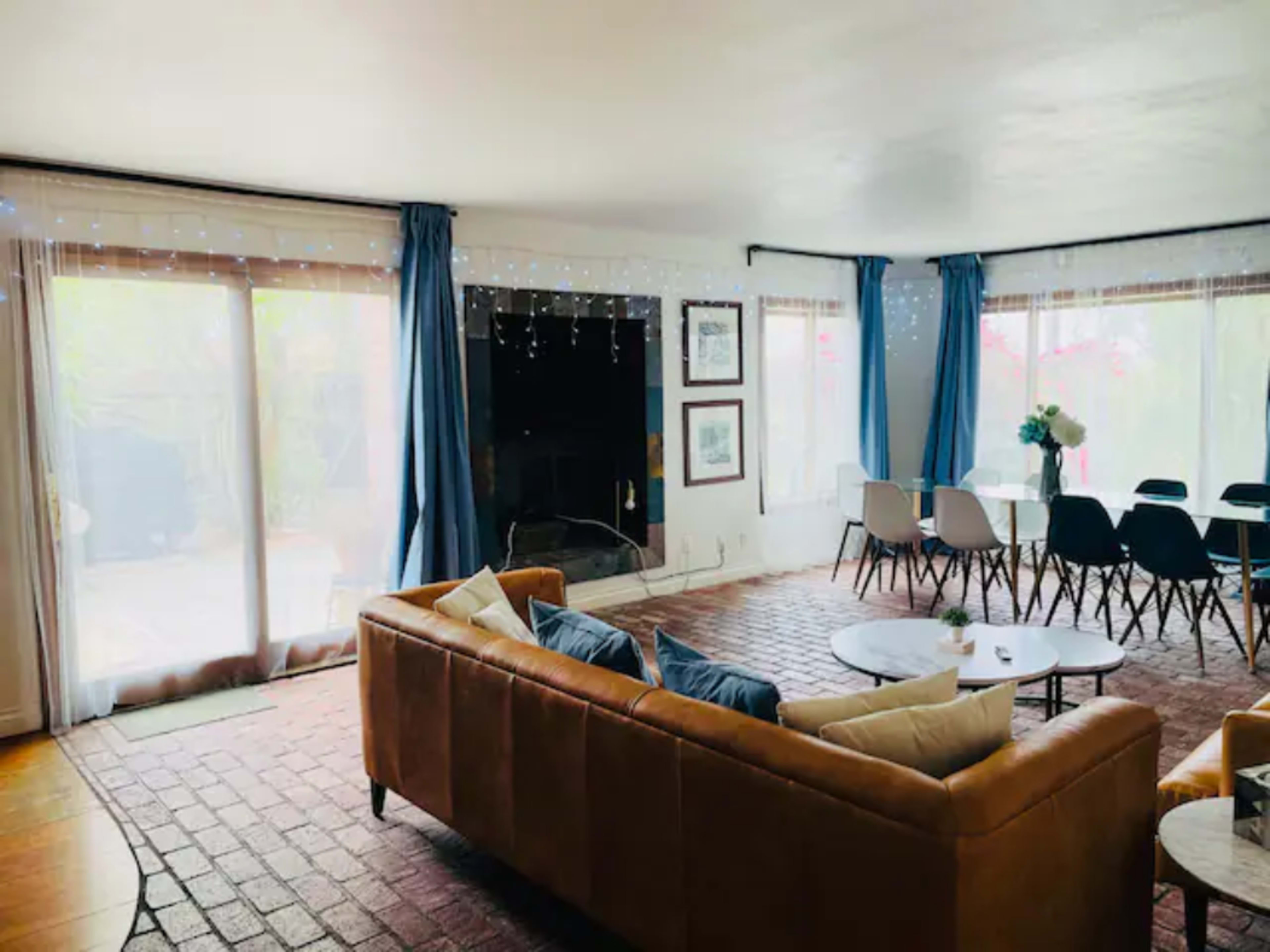 A spacious living area features a brown leather sofa, a round coffee table, and a dining area with white chairs beside large windows and sliding glass doors.