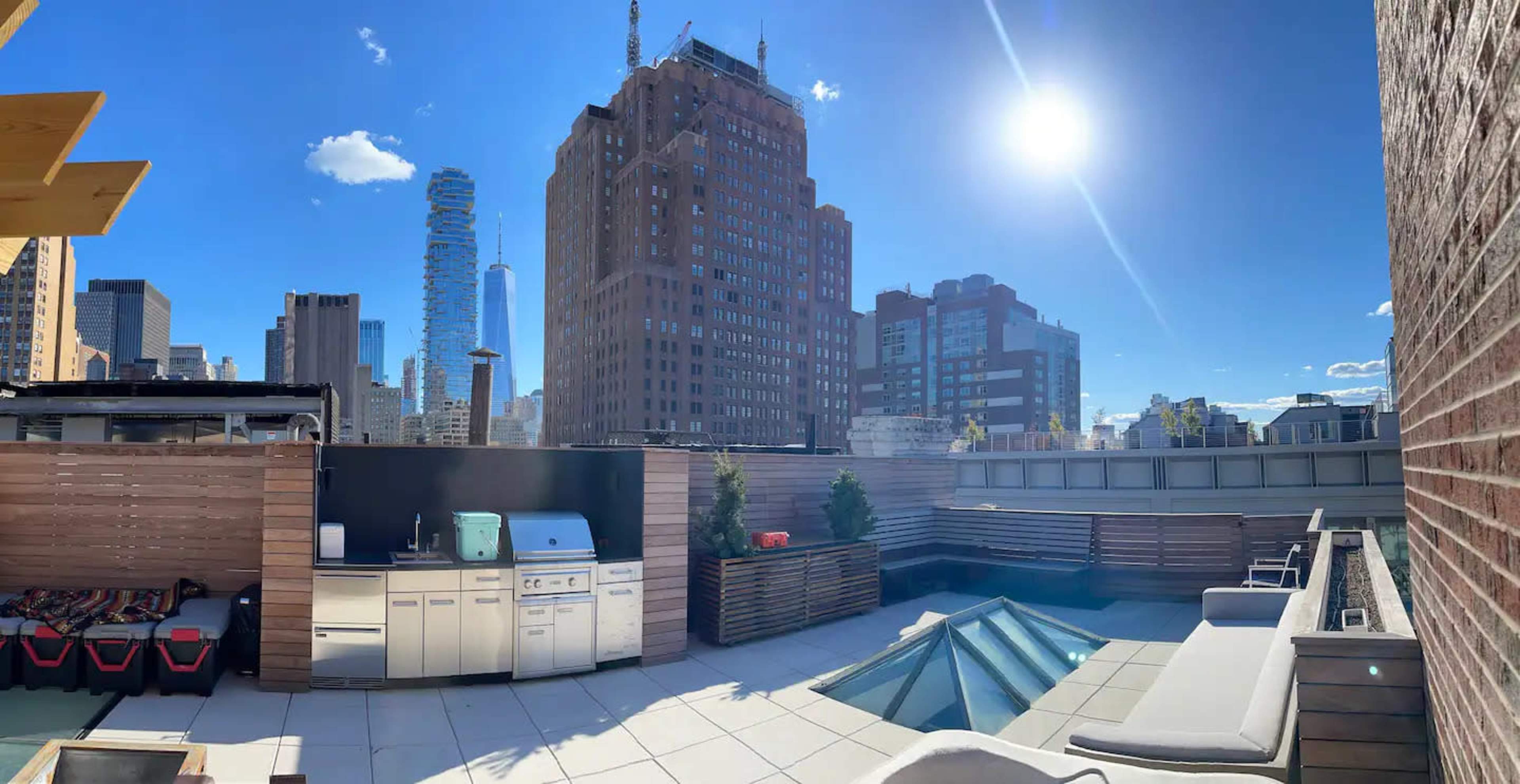 A rooftop terrace with an outdoor kitchen, seating area, and views of tall city buildings under a bright sky.