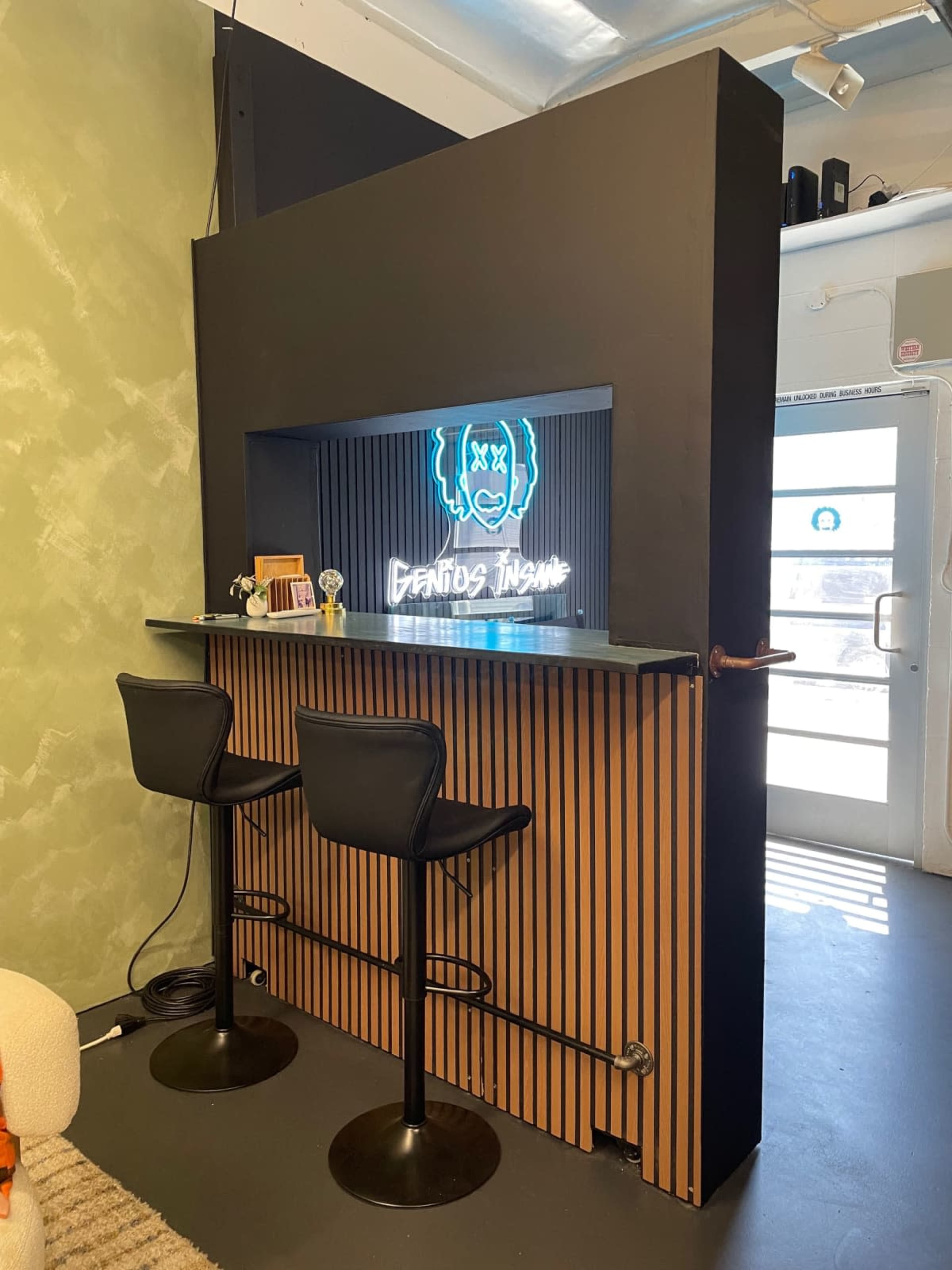 The image shows a modern bar area with two high stools, a neon sign that reads "DEALING INSANE," and a backdrop of vertical wooden slats.