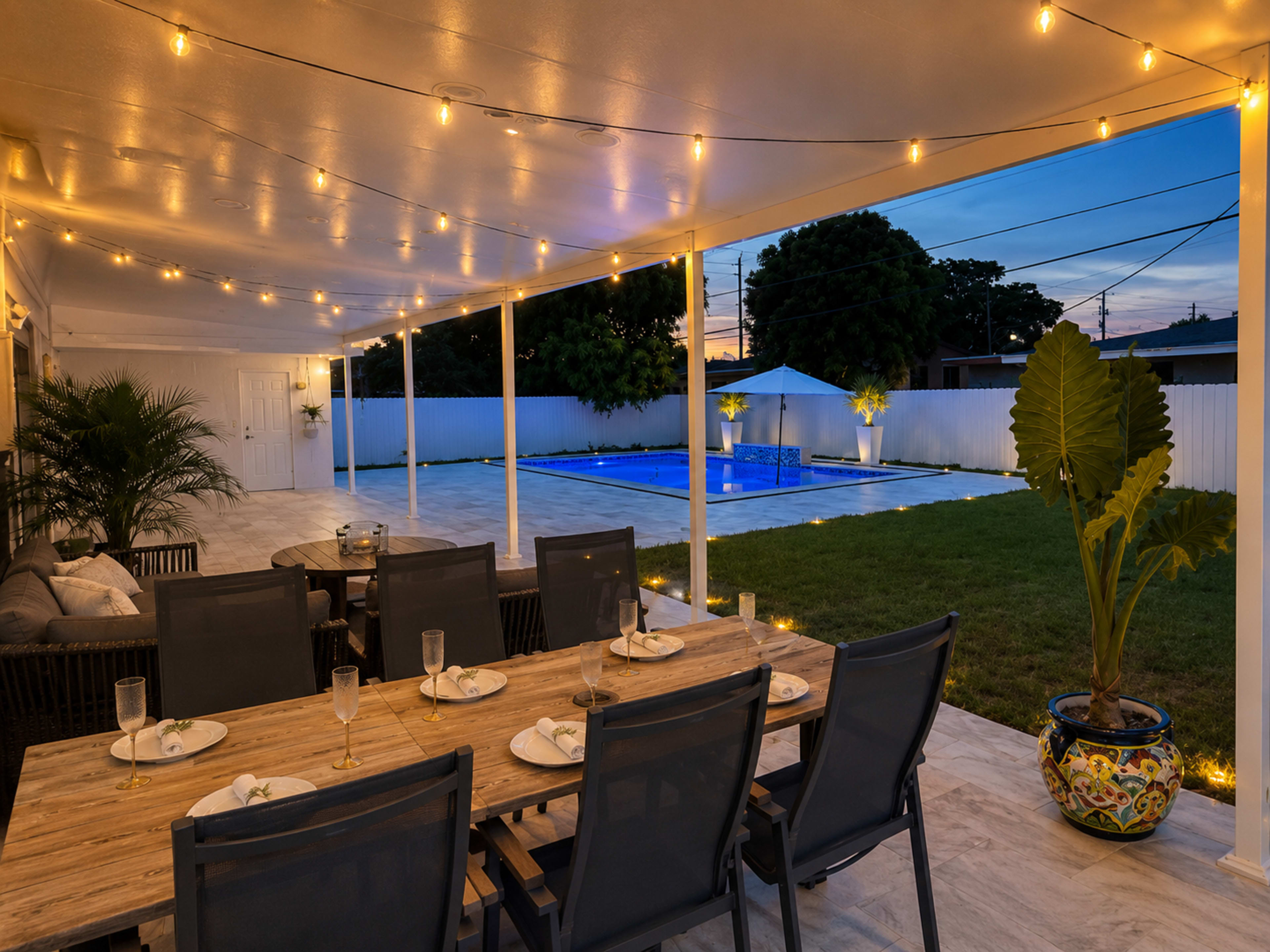 The image shows a covered outdoor dining area with a wooden table and chairs, overlooking a well-lit pool and a grassy yard at dusk.