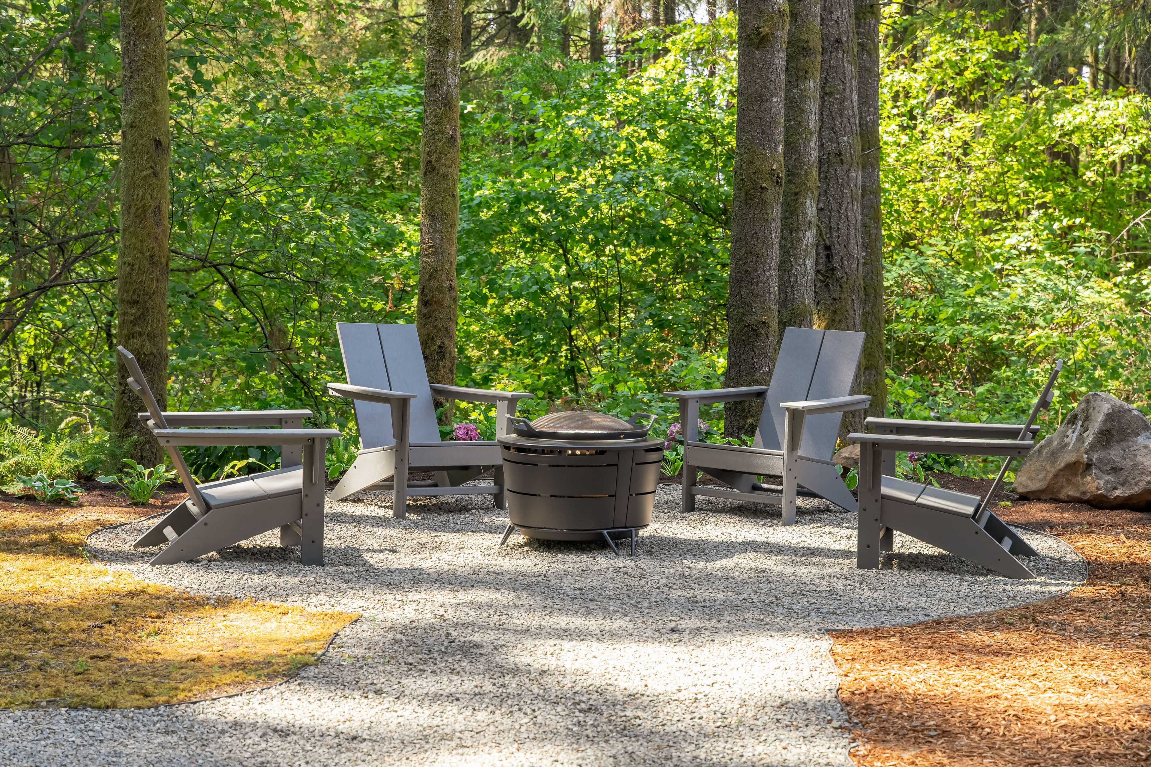 A circular seating arrangement with four gray chairs surrounds a fire pit on a gravel surface, set amidst trees and greenery.
