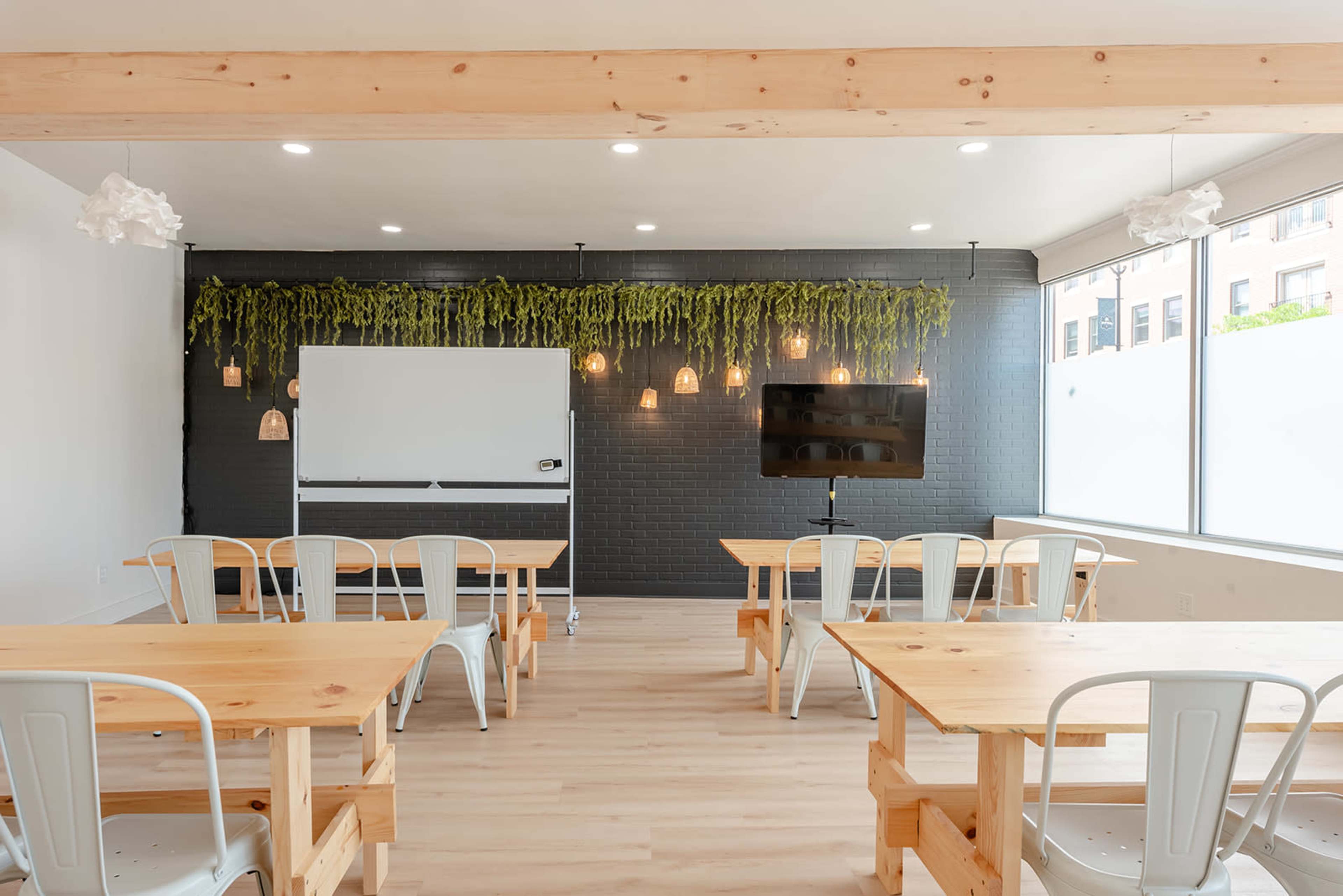 A bright meeting room features wooden tables and chairs, a whiteboard, a television, and greenery hanging from the walls.