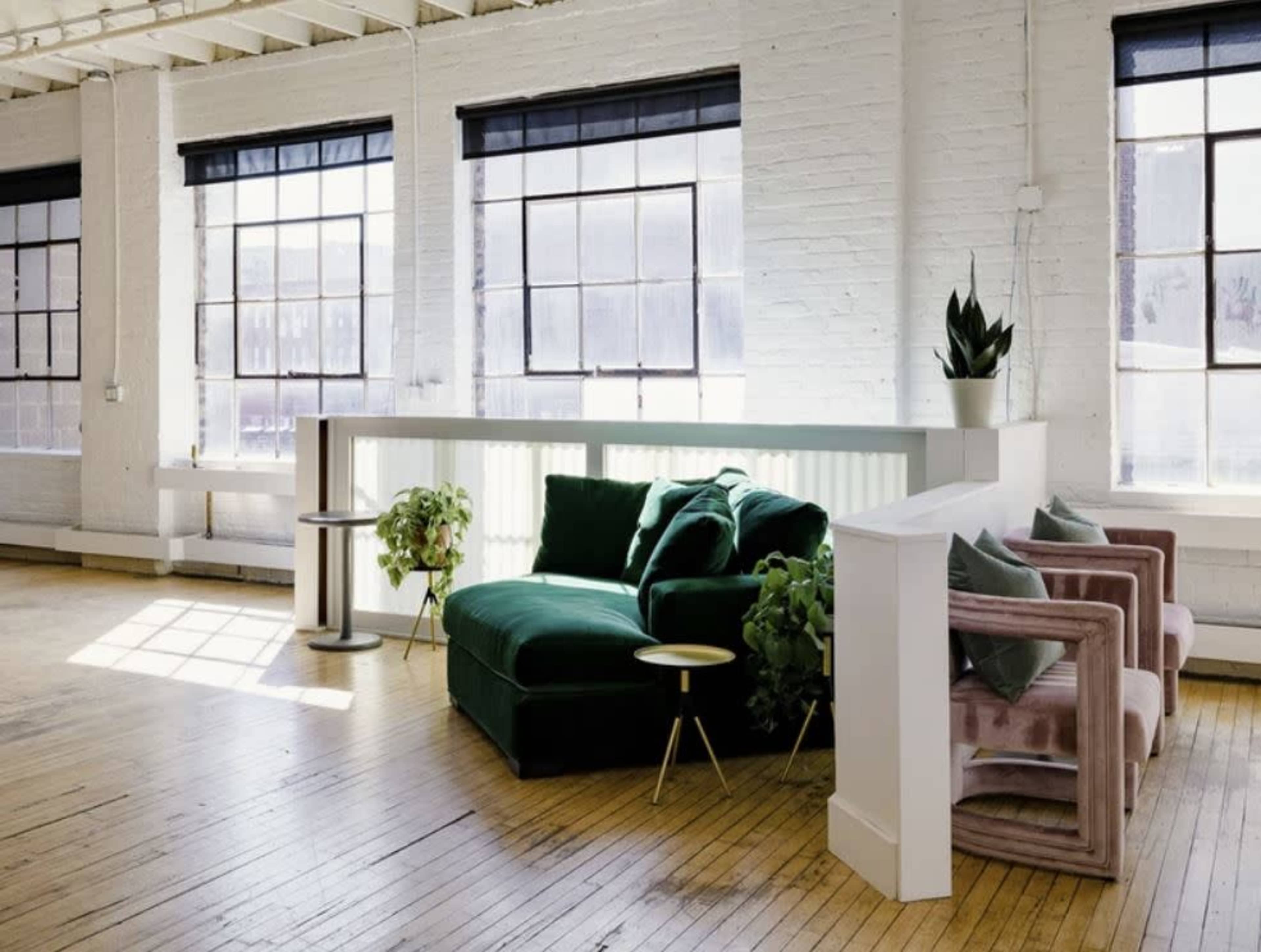 The image shows a modern, bright room with large windows, featuring a green sofa and pink chairs, surrounded by plants and wooden flooring.