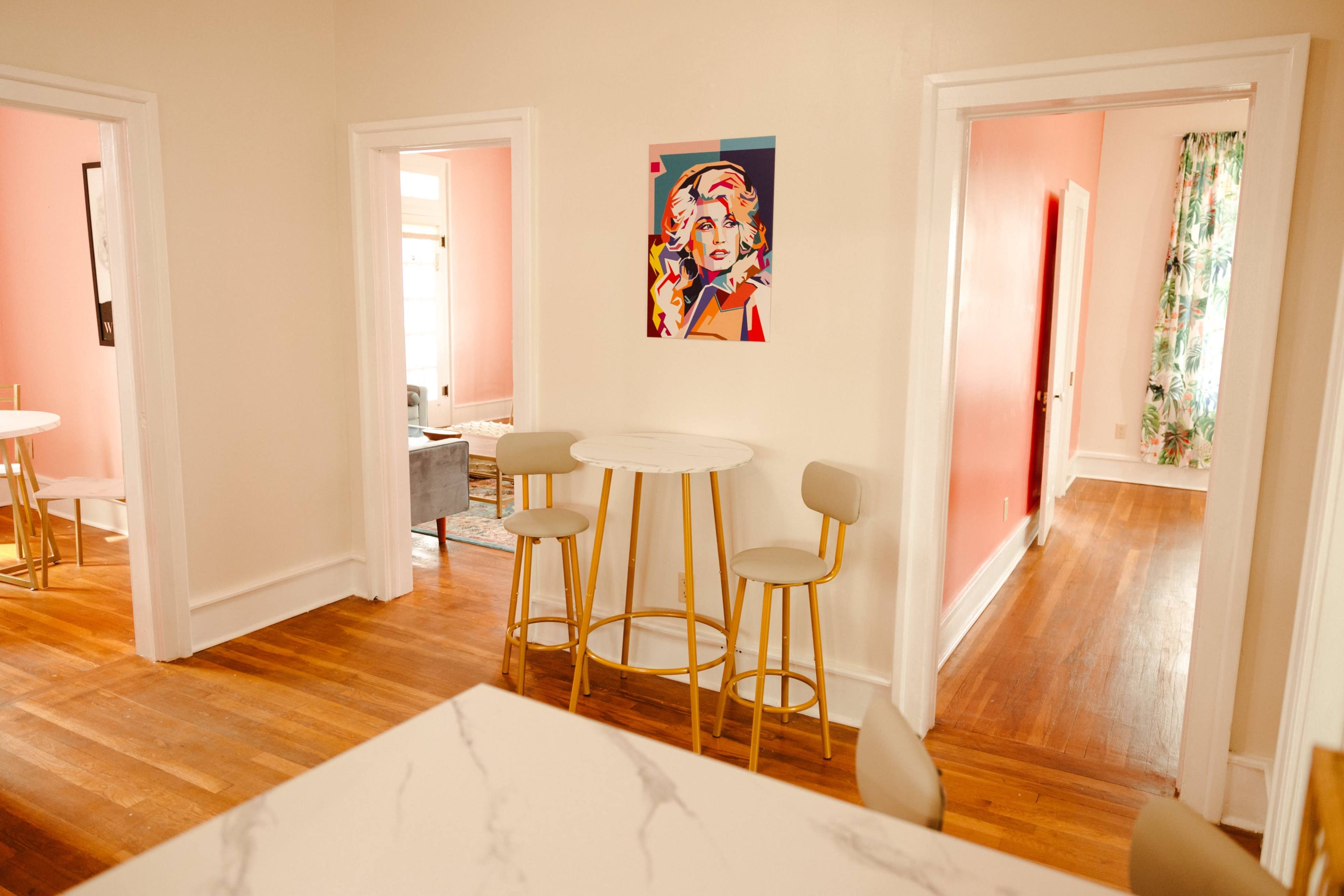 The image shows a bright room with a round table and stools, framed by doorways leading to adjacent rooms, featuring a colorful artwork on the wall.