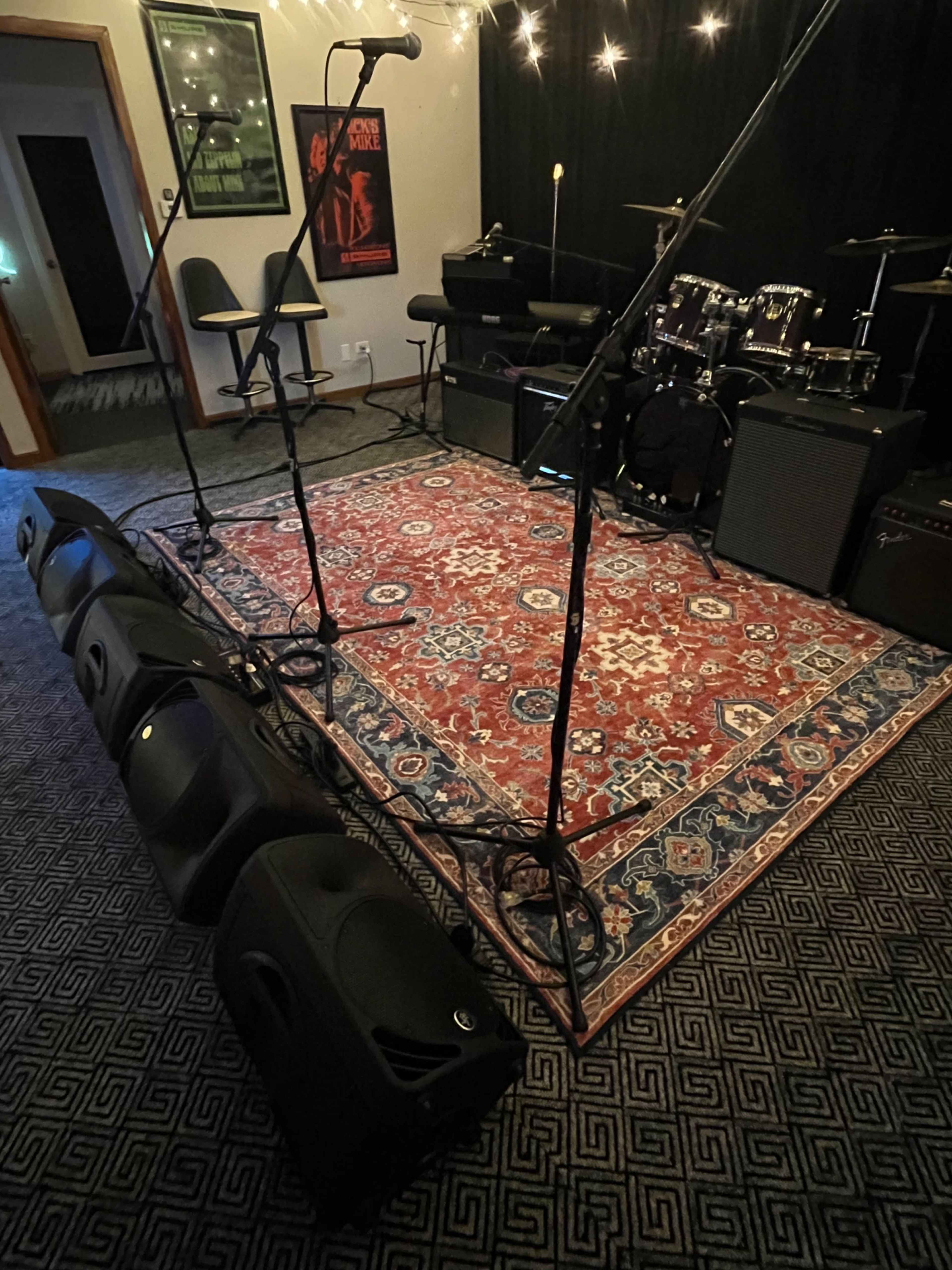 A stage setup features a red patterned rug, two microphone stands, and music equipment including drums and amplifiers in a dimly lit room.