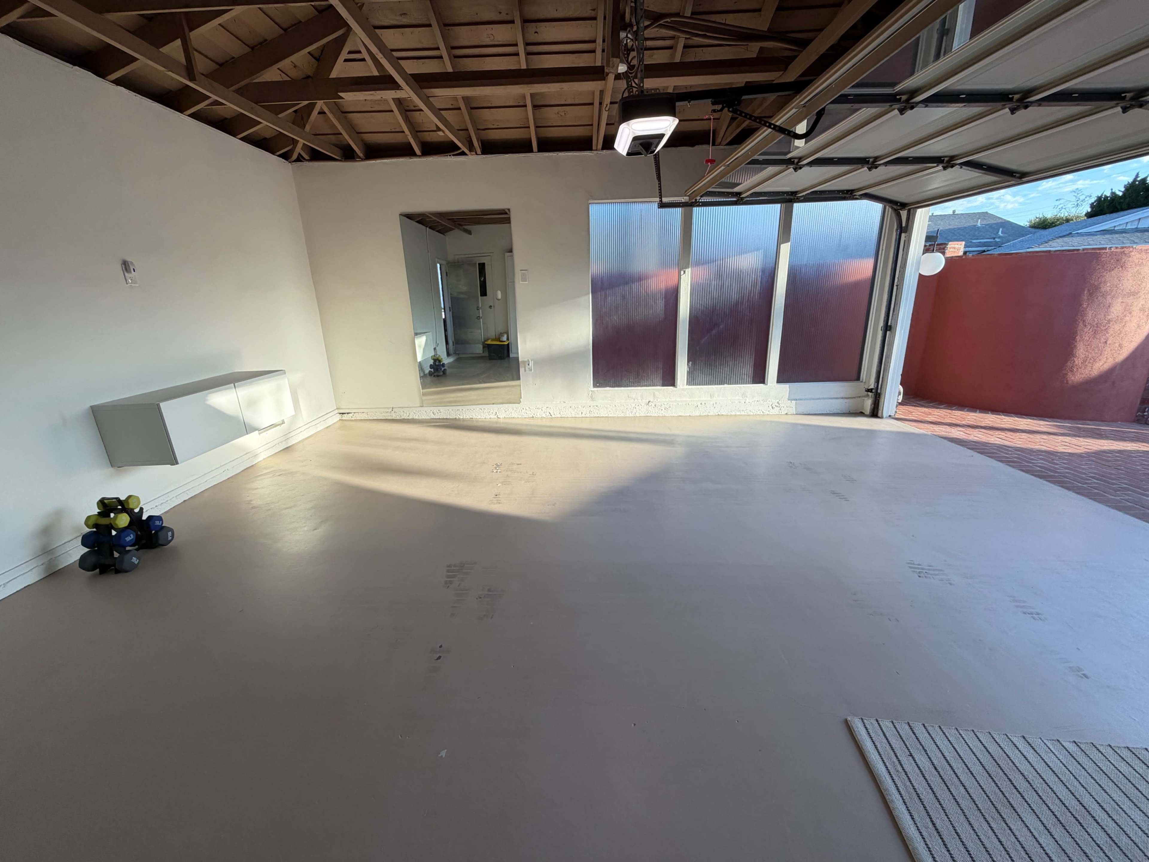 The image shows an empty garage with a clean floor, a small set of dumbbells in one corner, and sunlight coming through a large open door.