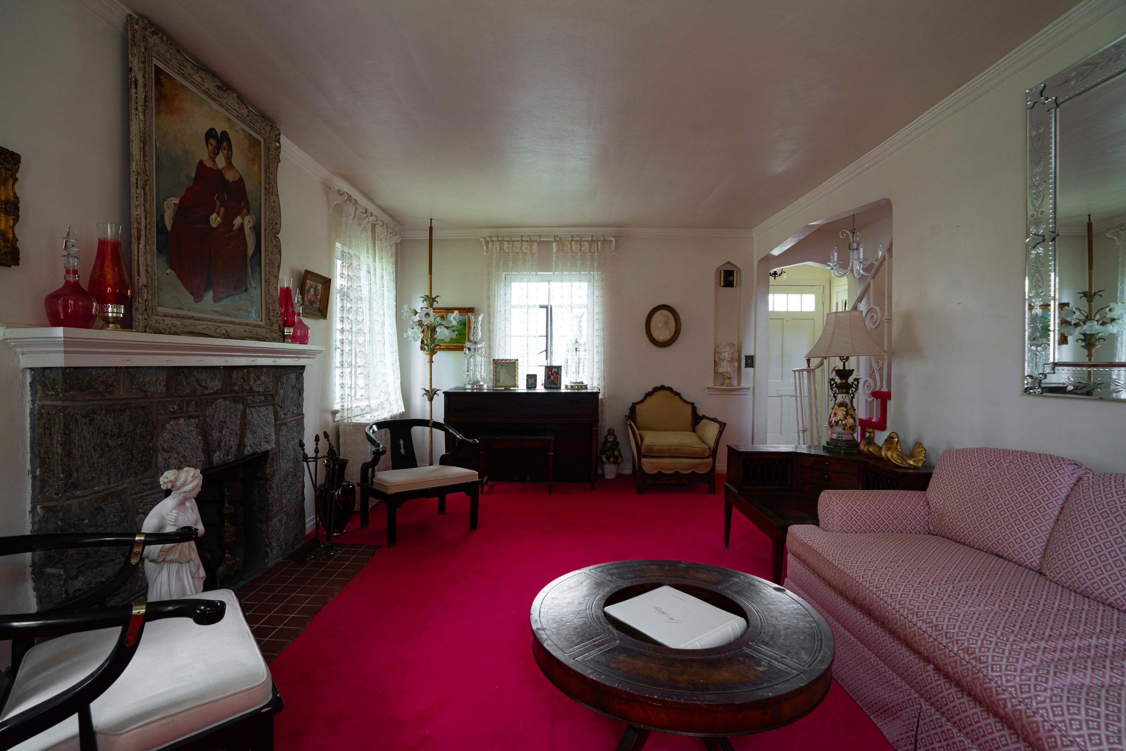 The living room features a pink carpet, a vintage sofa, and various decorative items, including a fireplace and framed artwork on the walls.
