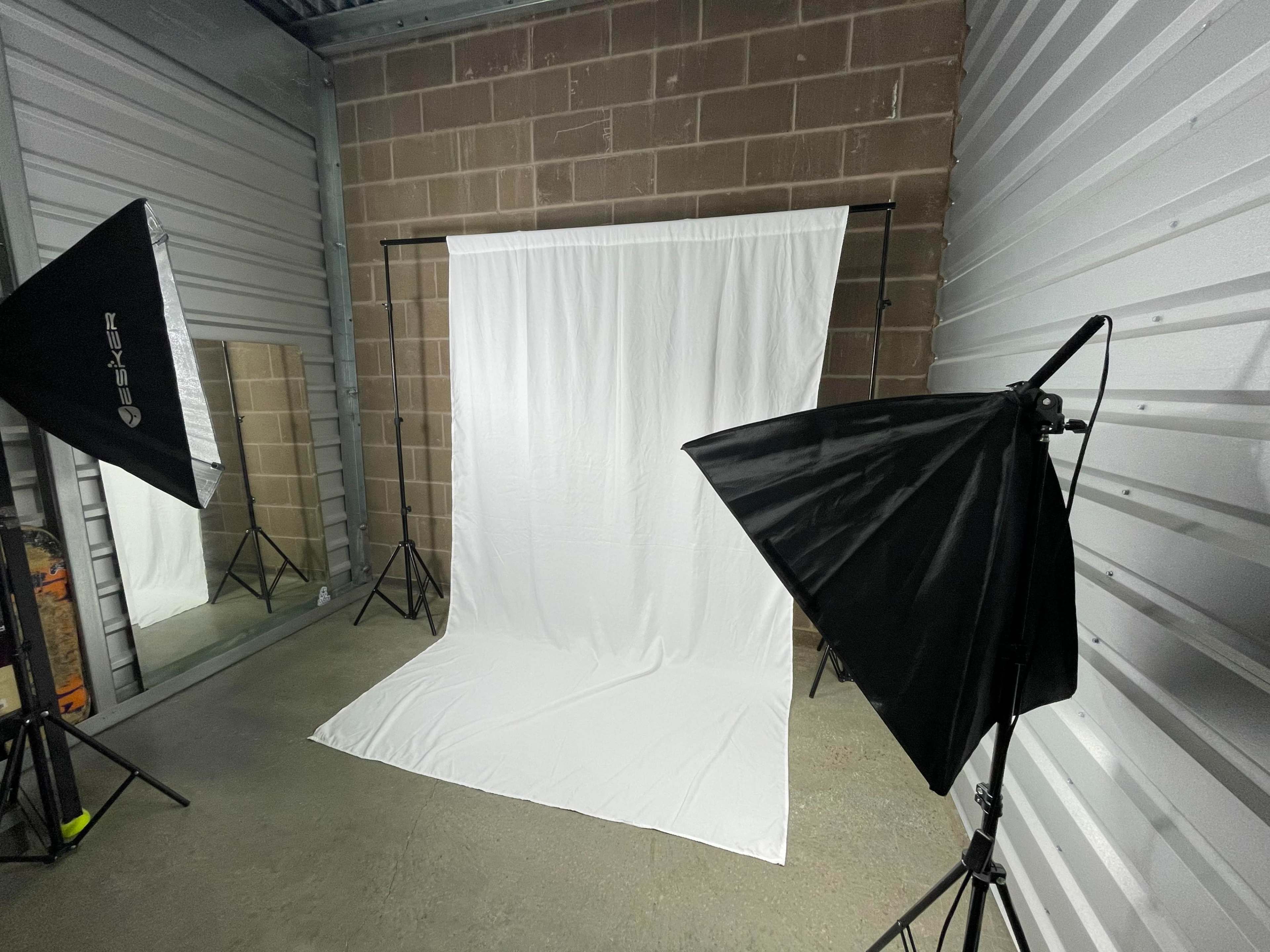 The image shows a photography studio setup with a white backdrop, two softbox lights, and a mirror along a concrete wall.