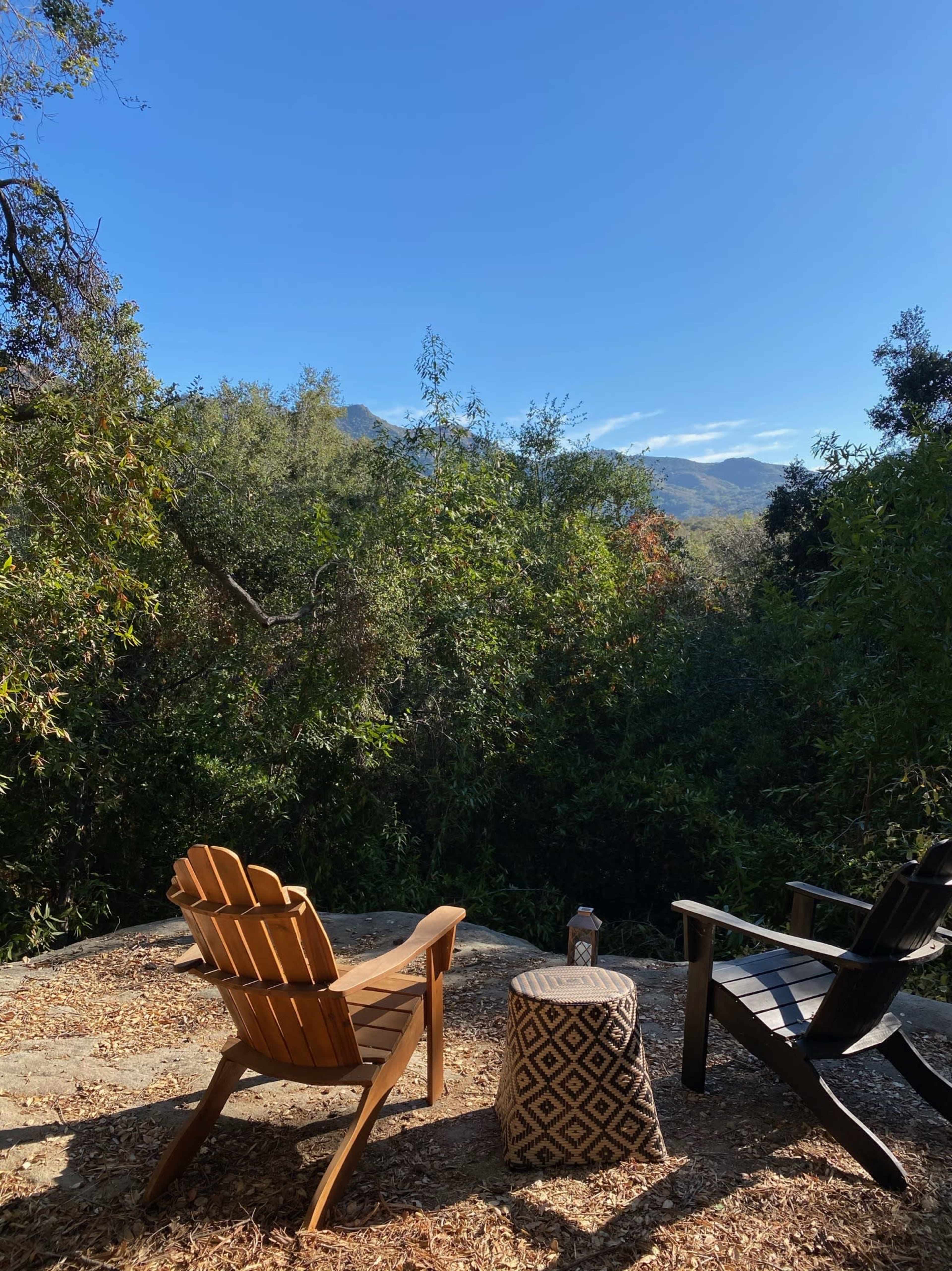 Two chairs sit on a stone patio surrounded by lush greenery, facing a scenic view of distant mountains under a clear blue sky.