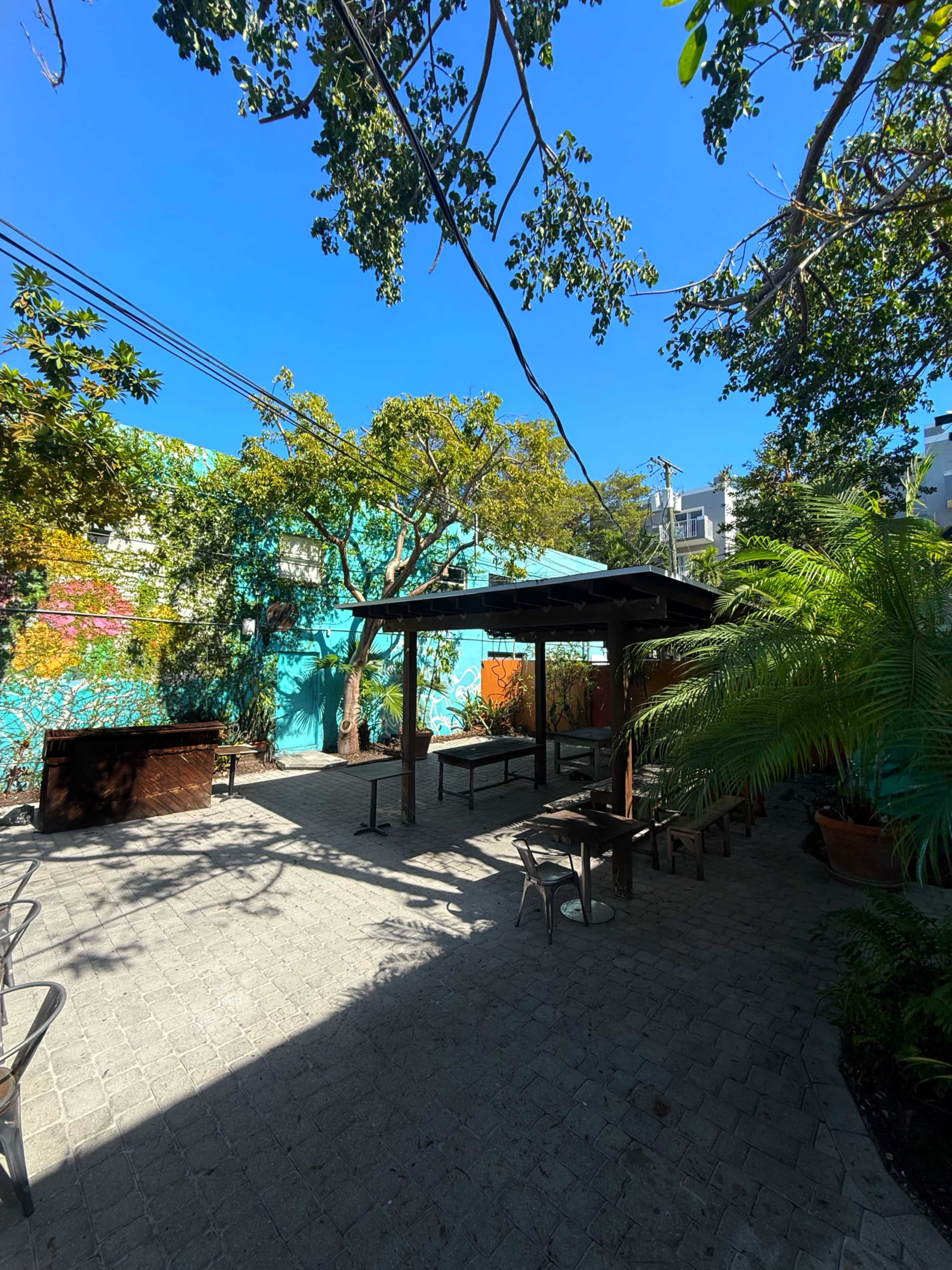 The scene features an outdoor patio area with a covered seating structure, surrounded by trees and a colorful mural on the wall.