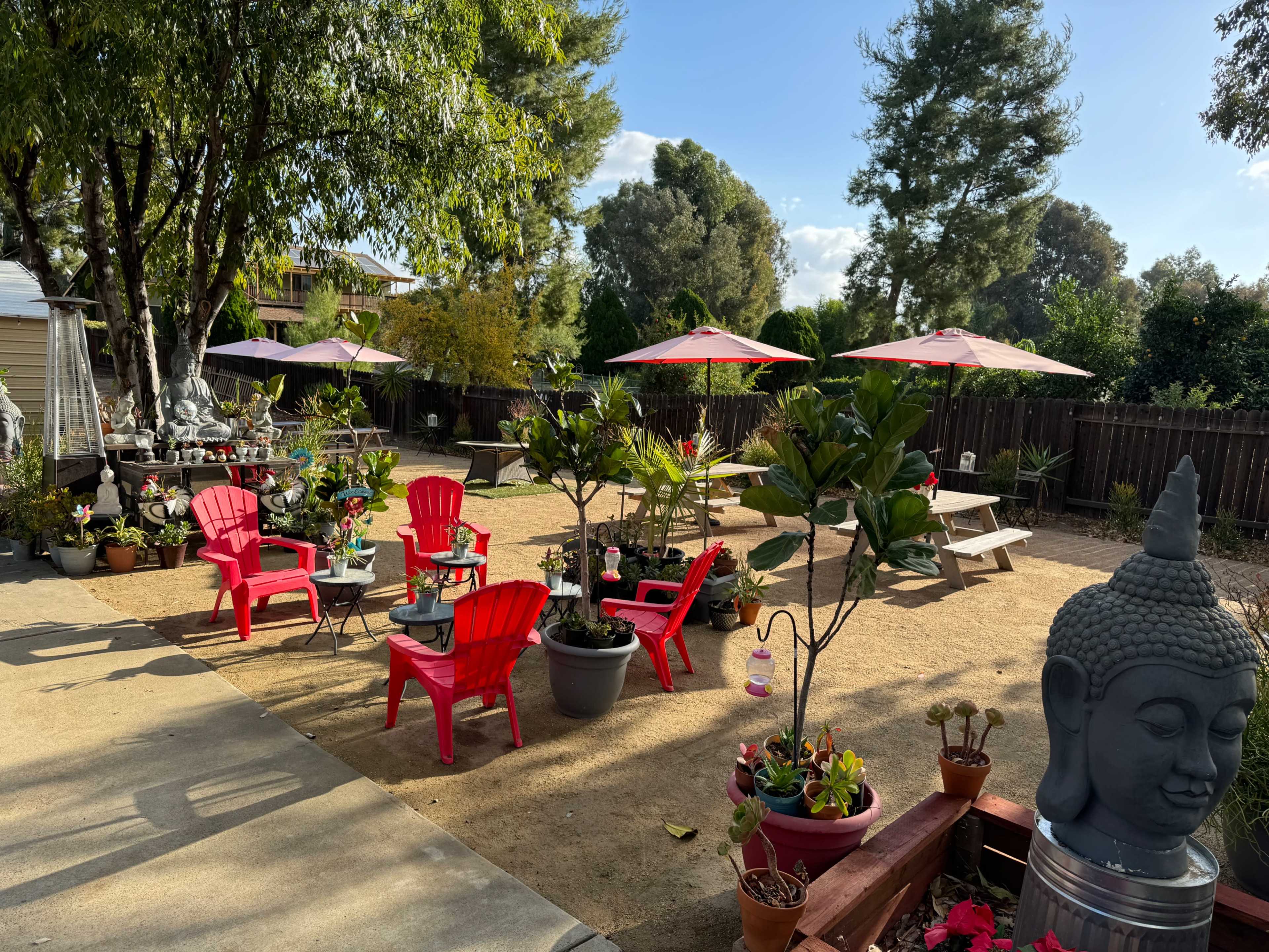 The image shows a landscaped garden area with red chairs, tables, umbrellas, and various potted plants, including a Buddha statue and seating arrangements.