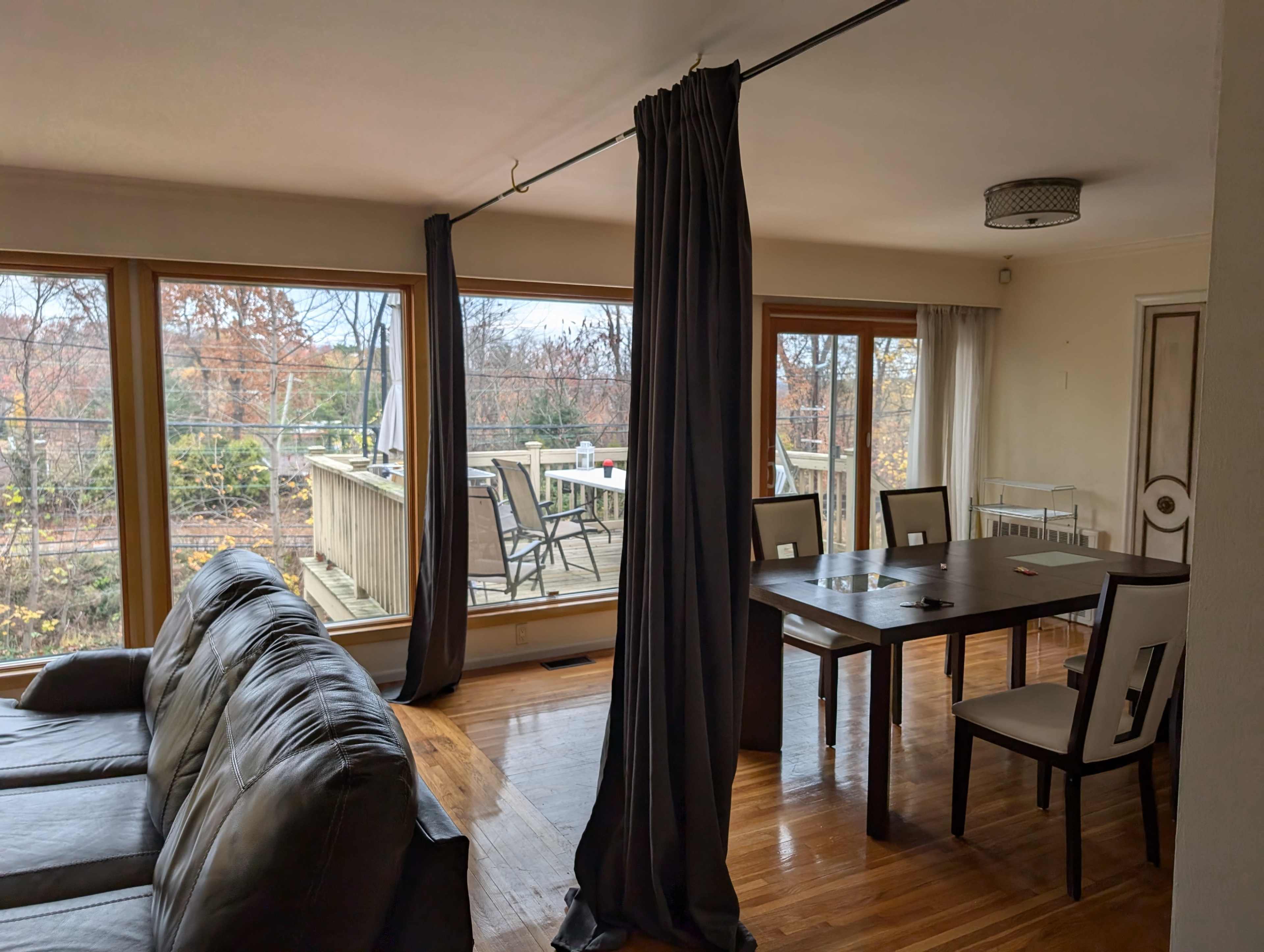 The image shows a living room with a sectional sofa, a dining table with chairs, and large windows overlooking a deck and outdoor area.