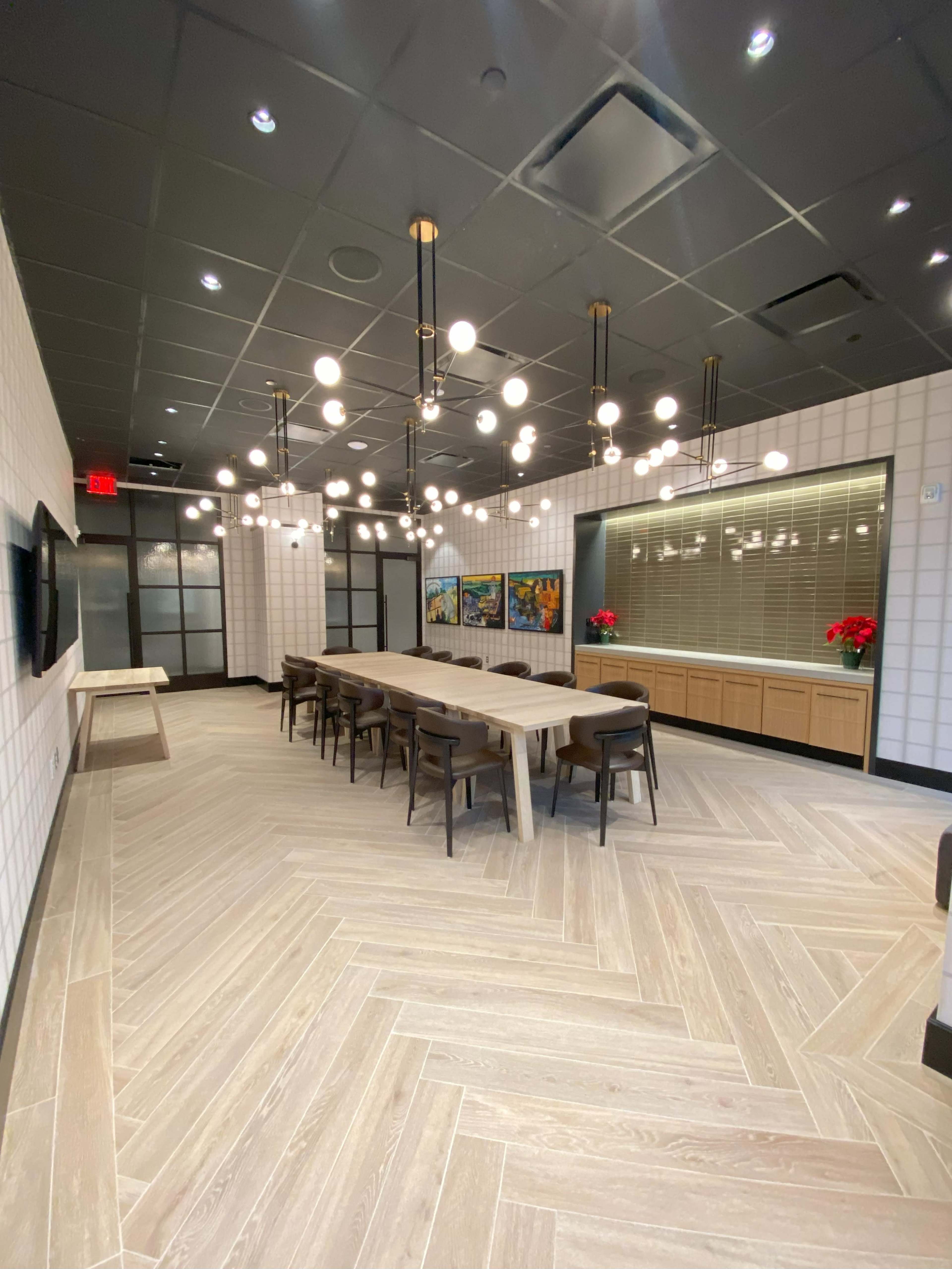 A modern conference room featuring a long wooden table surrounded by chairs, illuminated by spherical pendant lights, with decorative art and potted plants along the walls.
