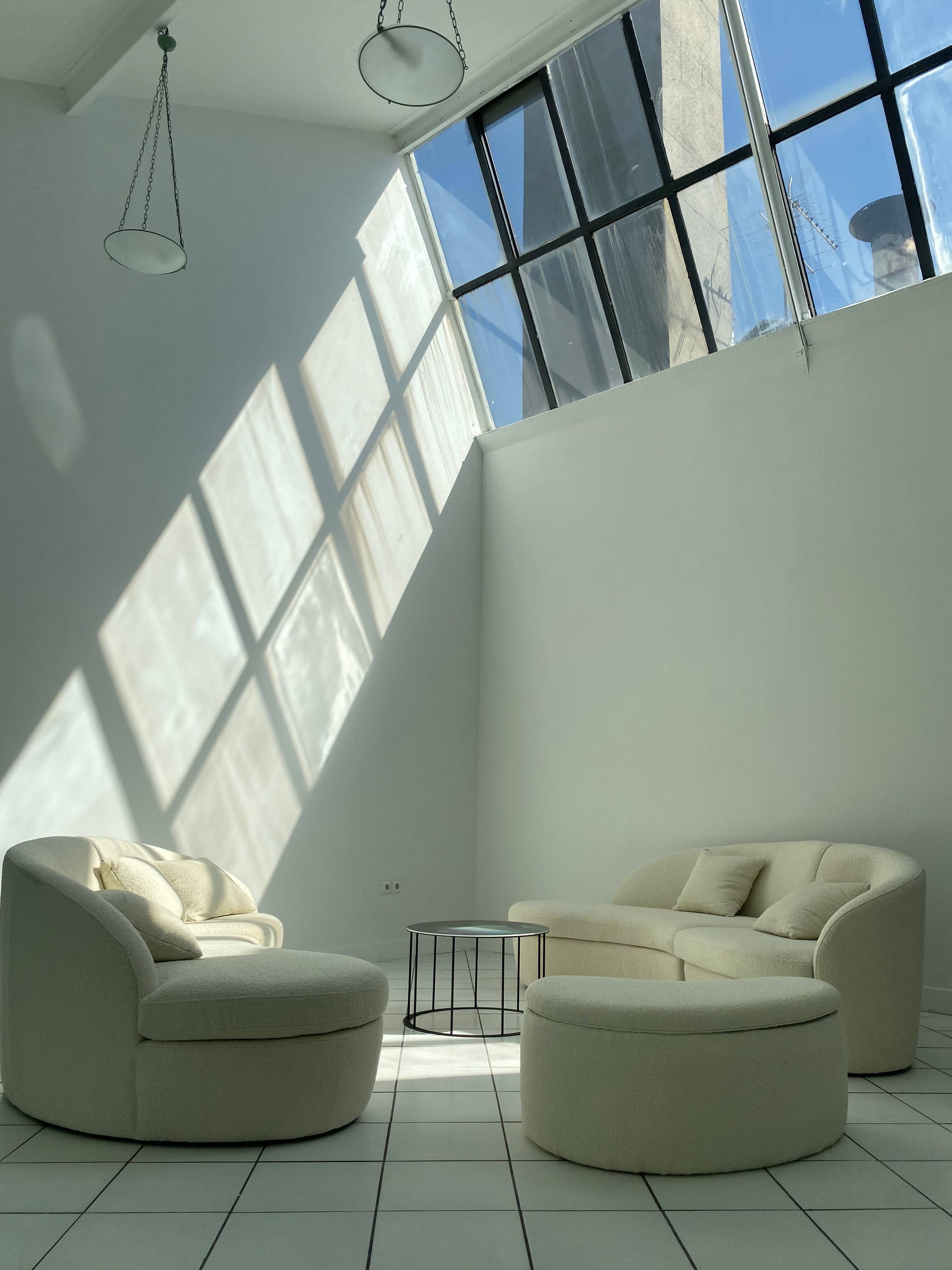 A minimalist living space with white furniture and large windows casting shadows on the tiled floor.