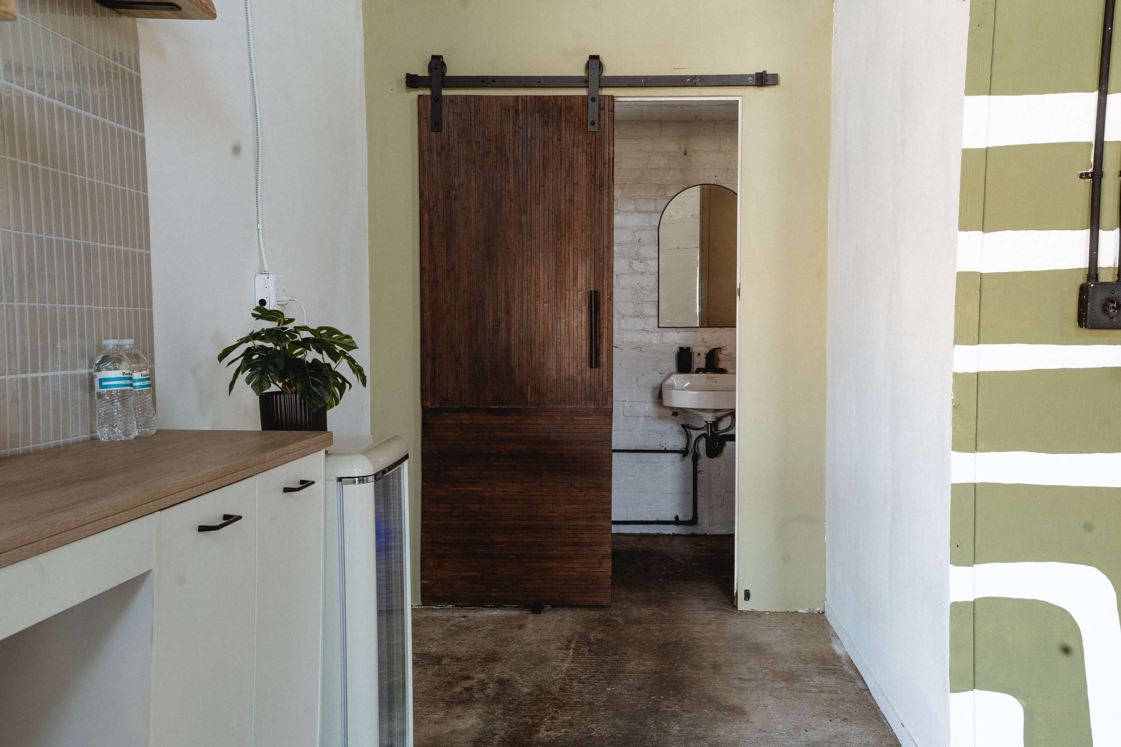 A sliding wooden door leads to a bathroom area with a sink and mirror, while a small plant and a bottle of water sit on a counter nearby.