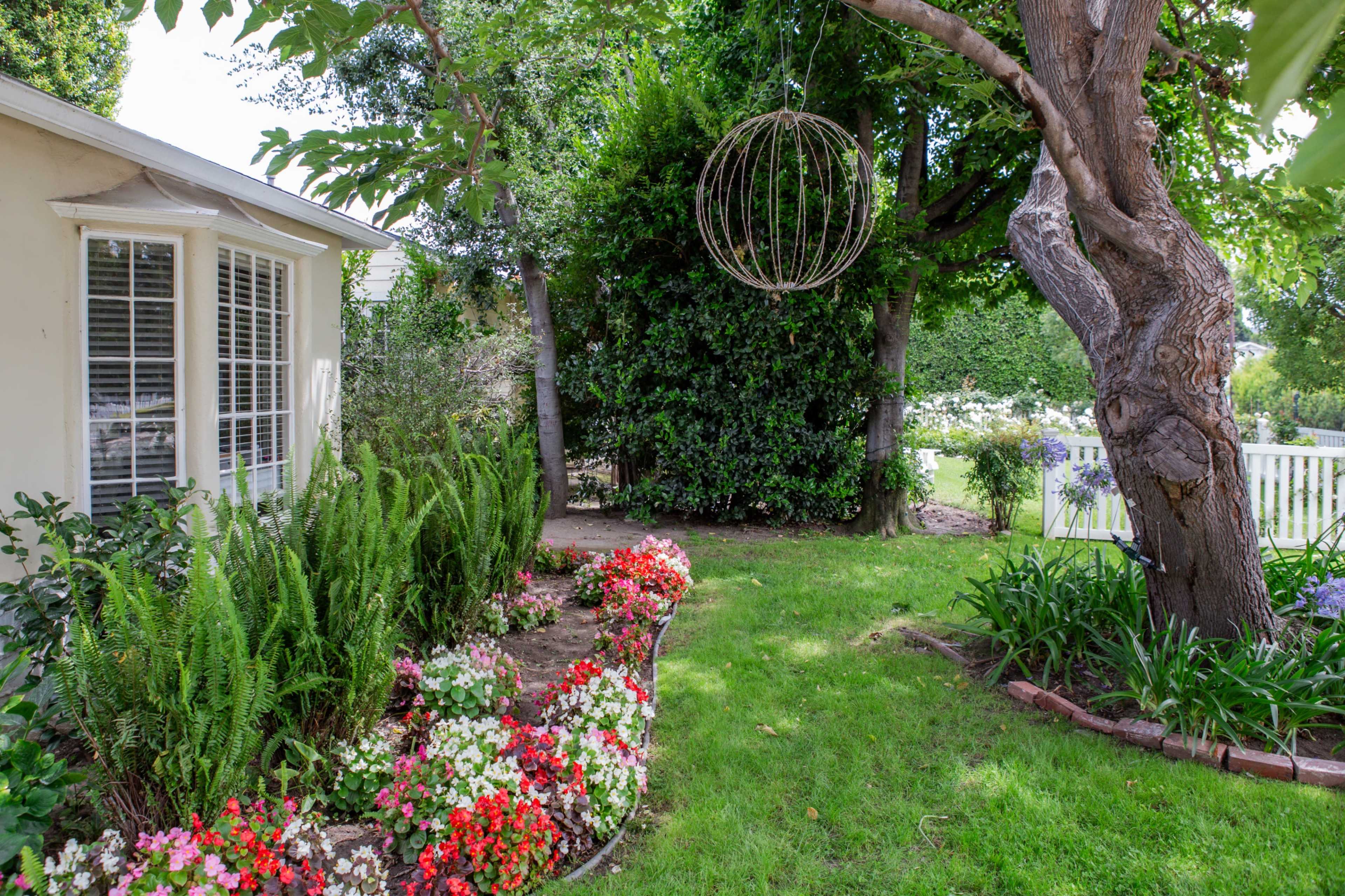 A garden scene features a curved flower bed with colorful flowers in front of a house, alongside a large tree adorned with a decorative hanging orb.
