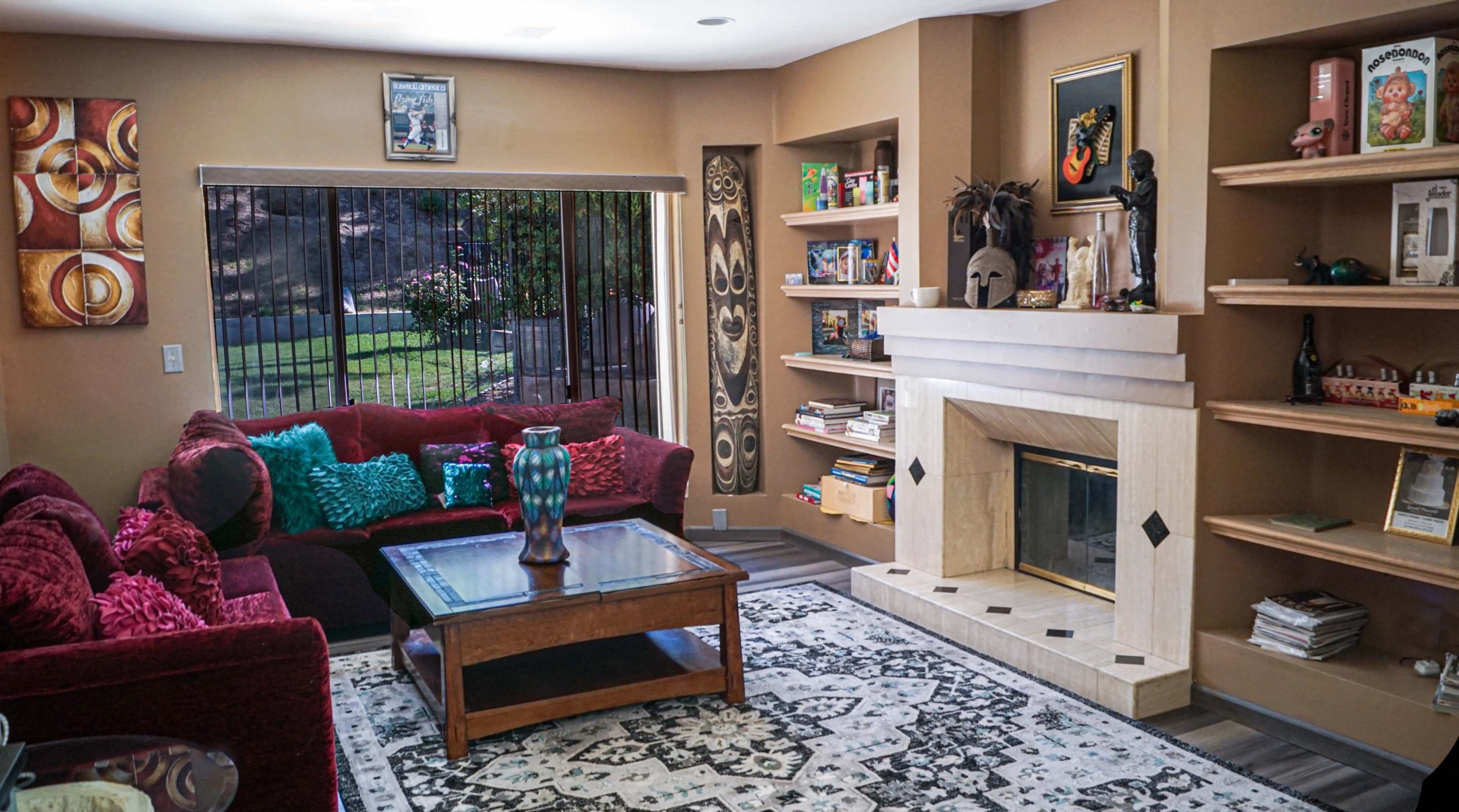 The image shows a living room featuring a red couch, a wooden coffee table, a fireplace with a decorative facade, and shelves filled with books and decorative items, all situated near a large window with a view of the outdoors.