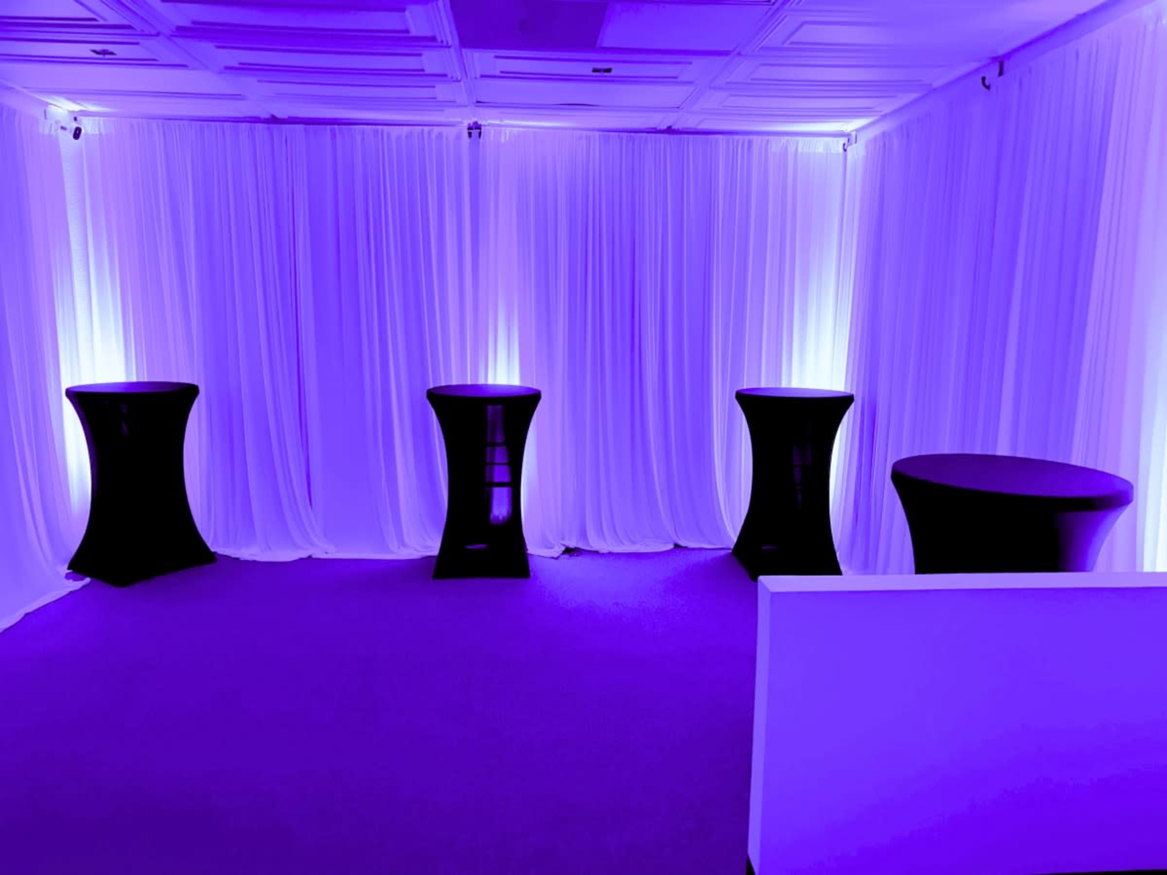 The image shows a room with four black cocktail tables illuminated by soft purple lighting against a backdrop of white drapery.