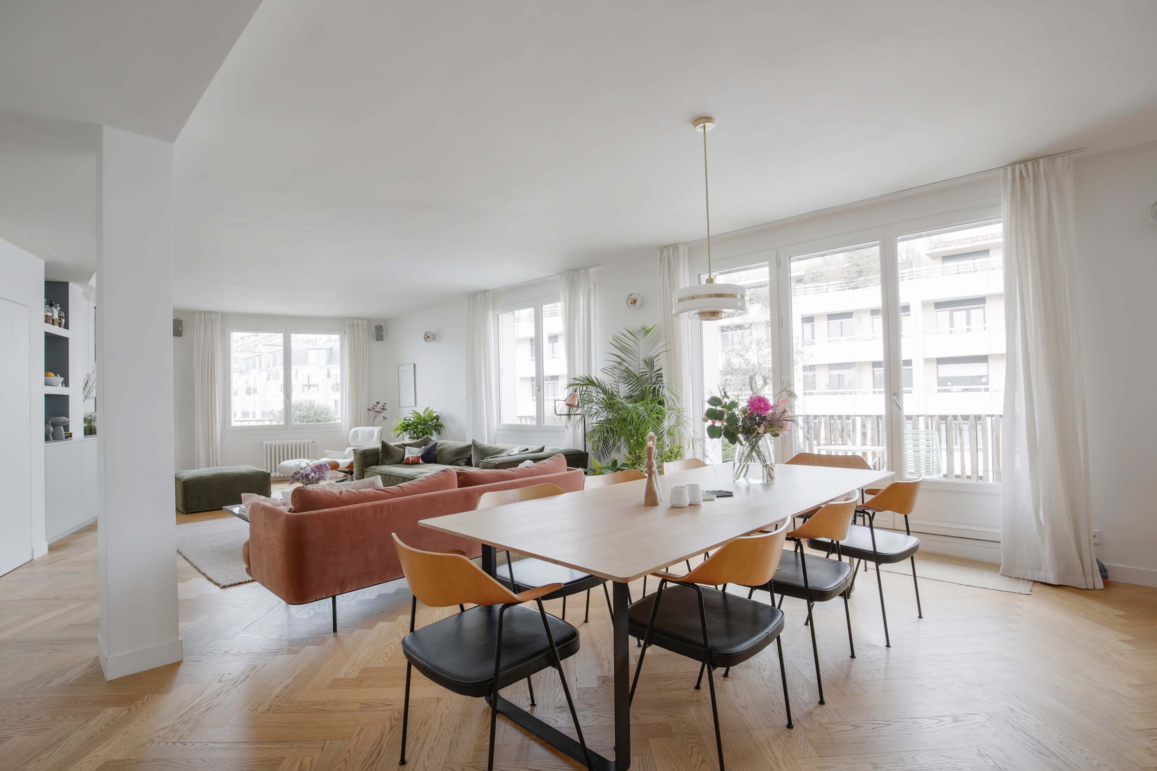 A modern, open-concept living and dining area with large windows, a wooden dining table, and a mix of green and orange seating.