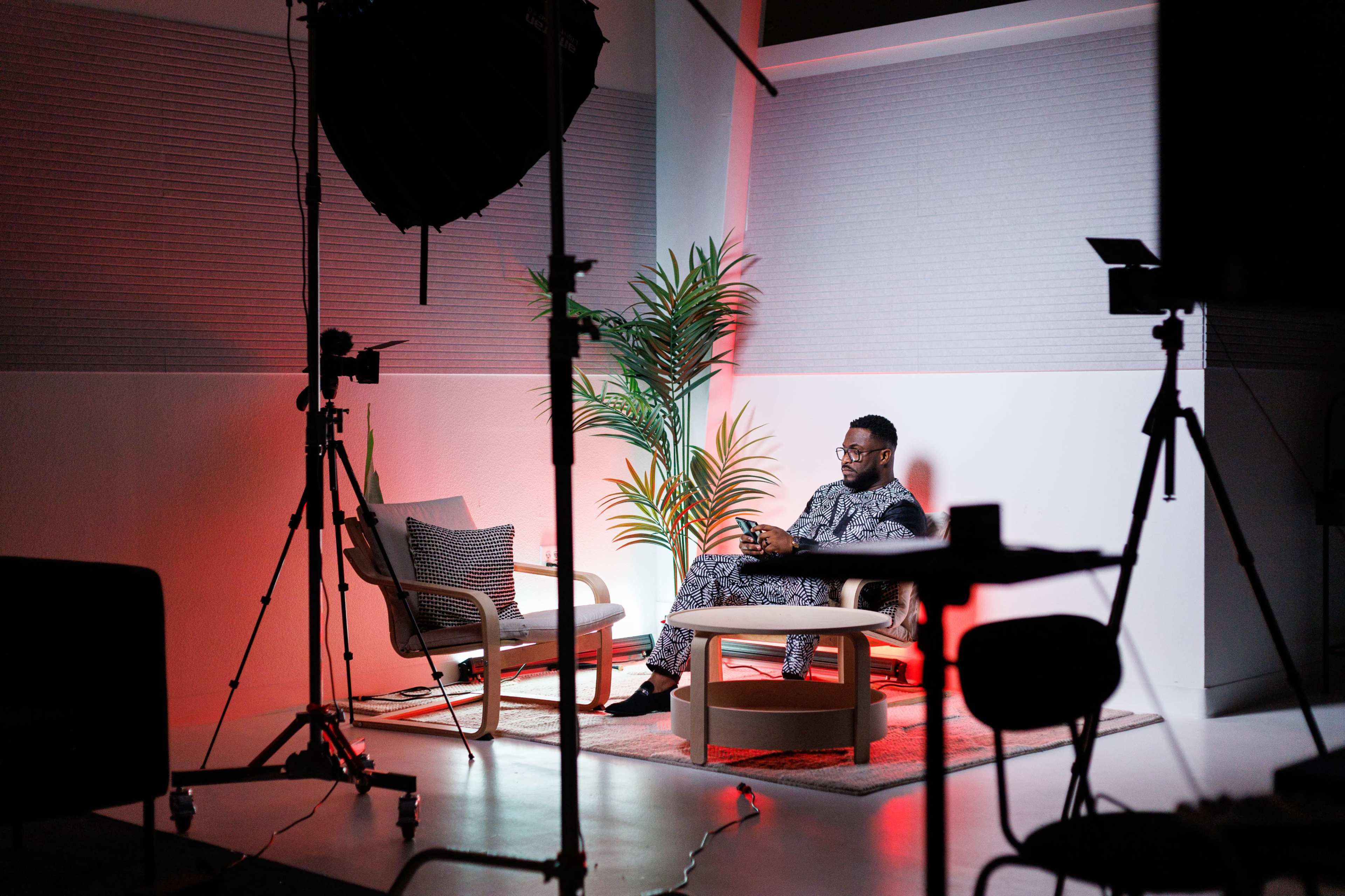 A person sits on a couch in a well-lit studio surrounded by cameras and lighting equipment.