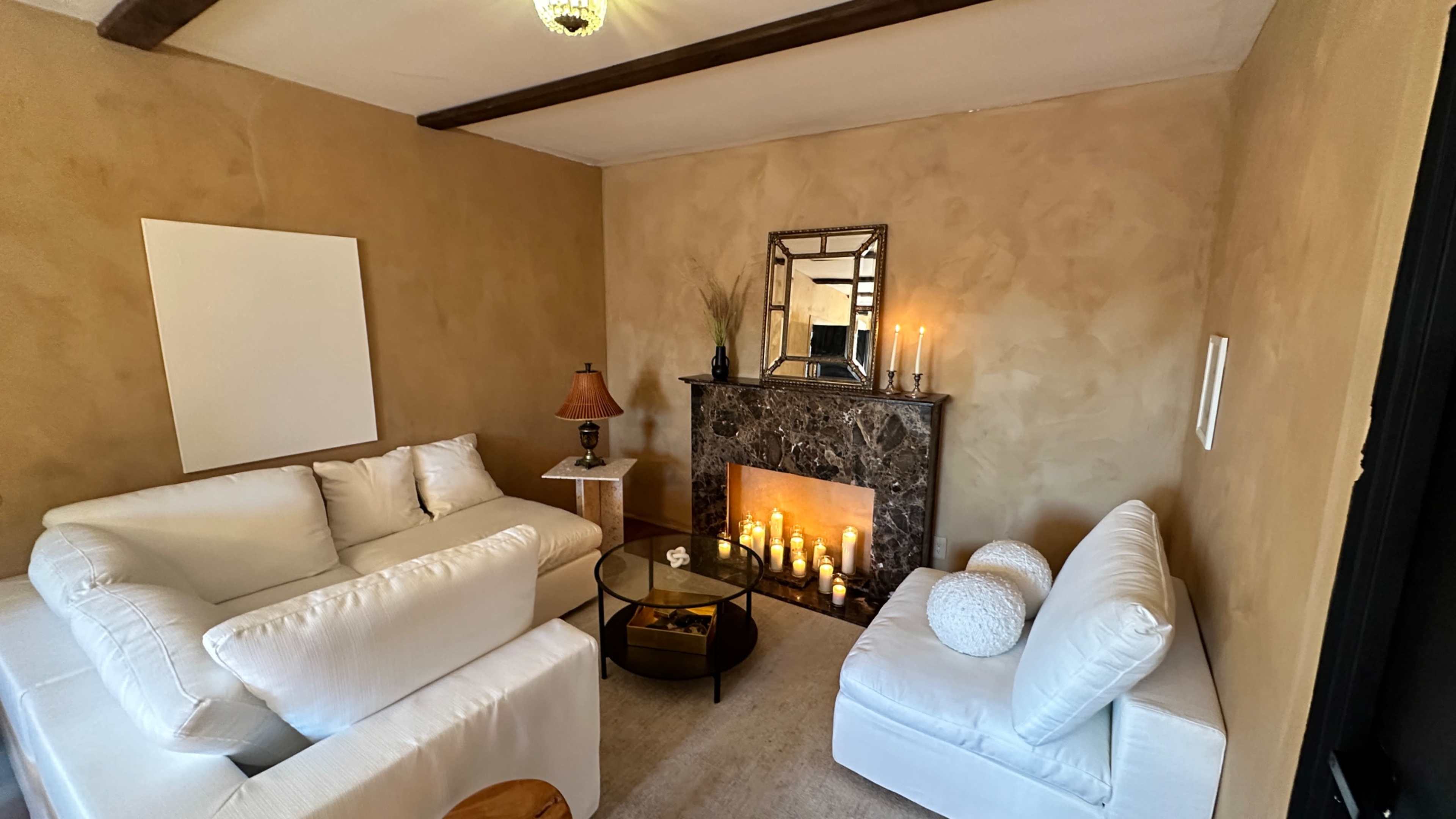 A cozy living room featuring white sofas, a marble fireplace with candles, and a small glass coffee table.