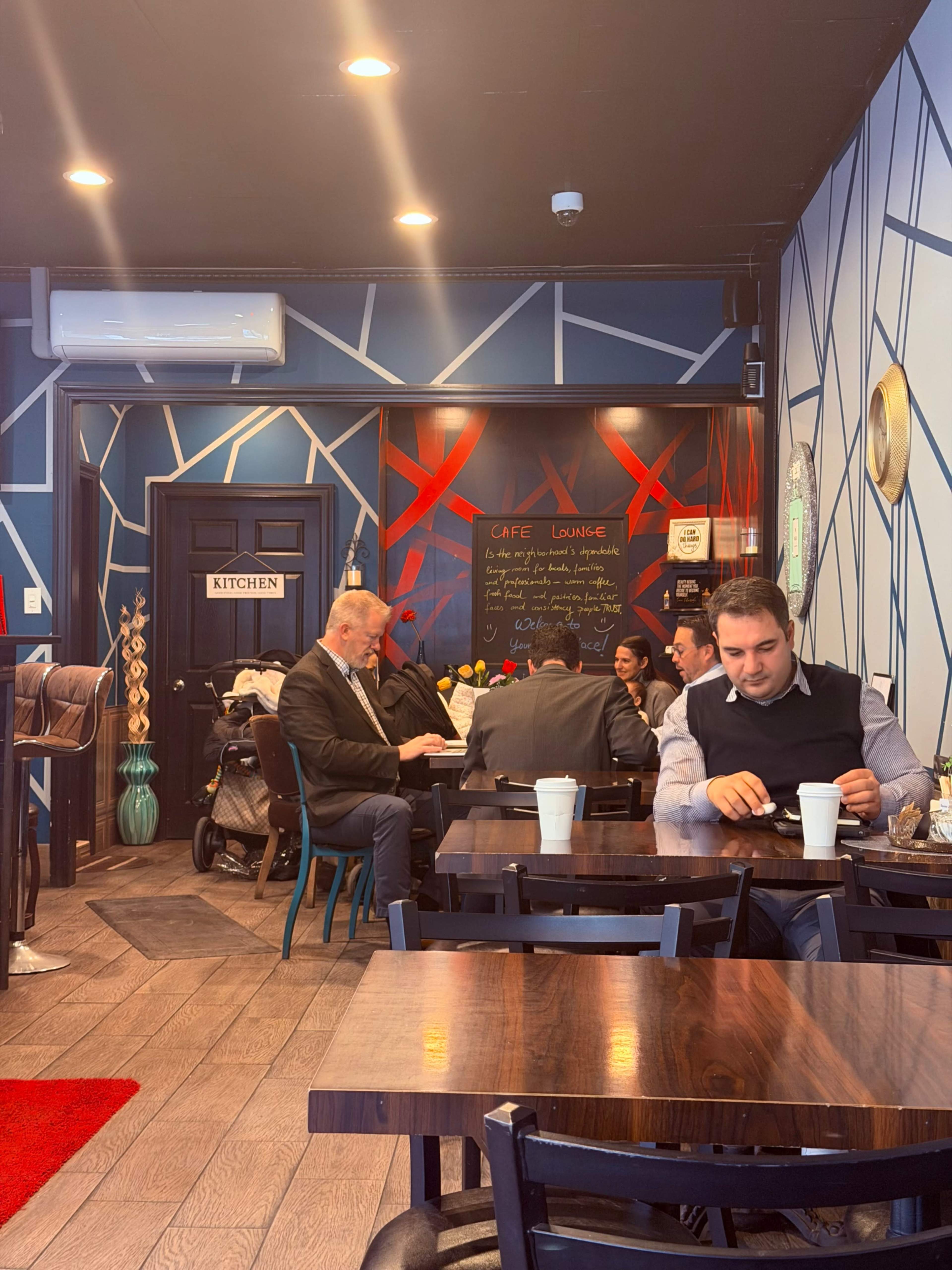 A group of men in business attire sits at a table in a brightly decorated café while one man drinks coffee alone at another table.