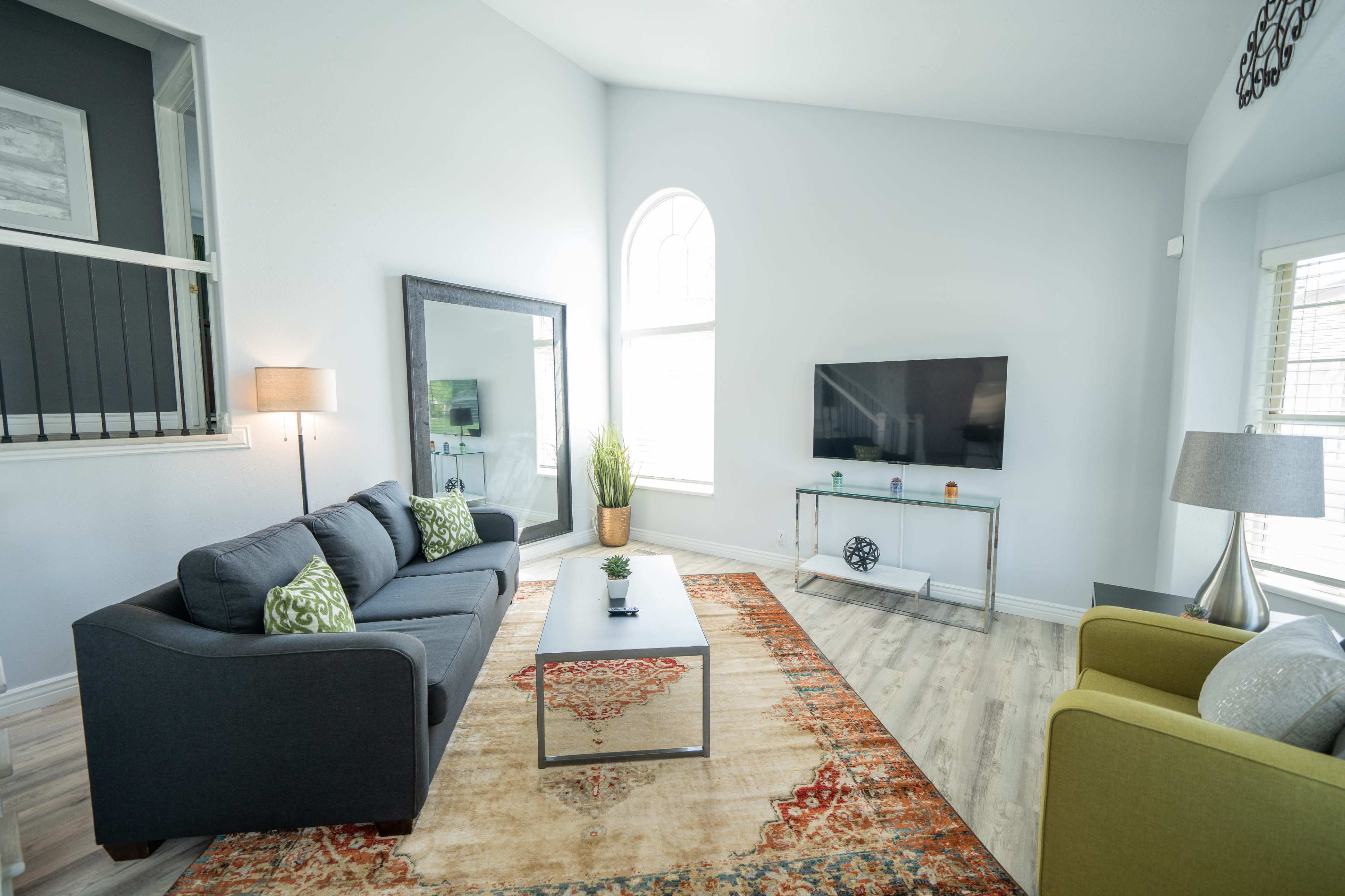 The room features a modern living area with a gray sofa, a green chair, a glass TV stand, and large windows allowing natural light.