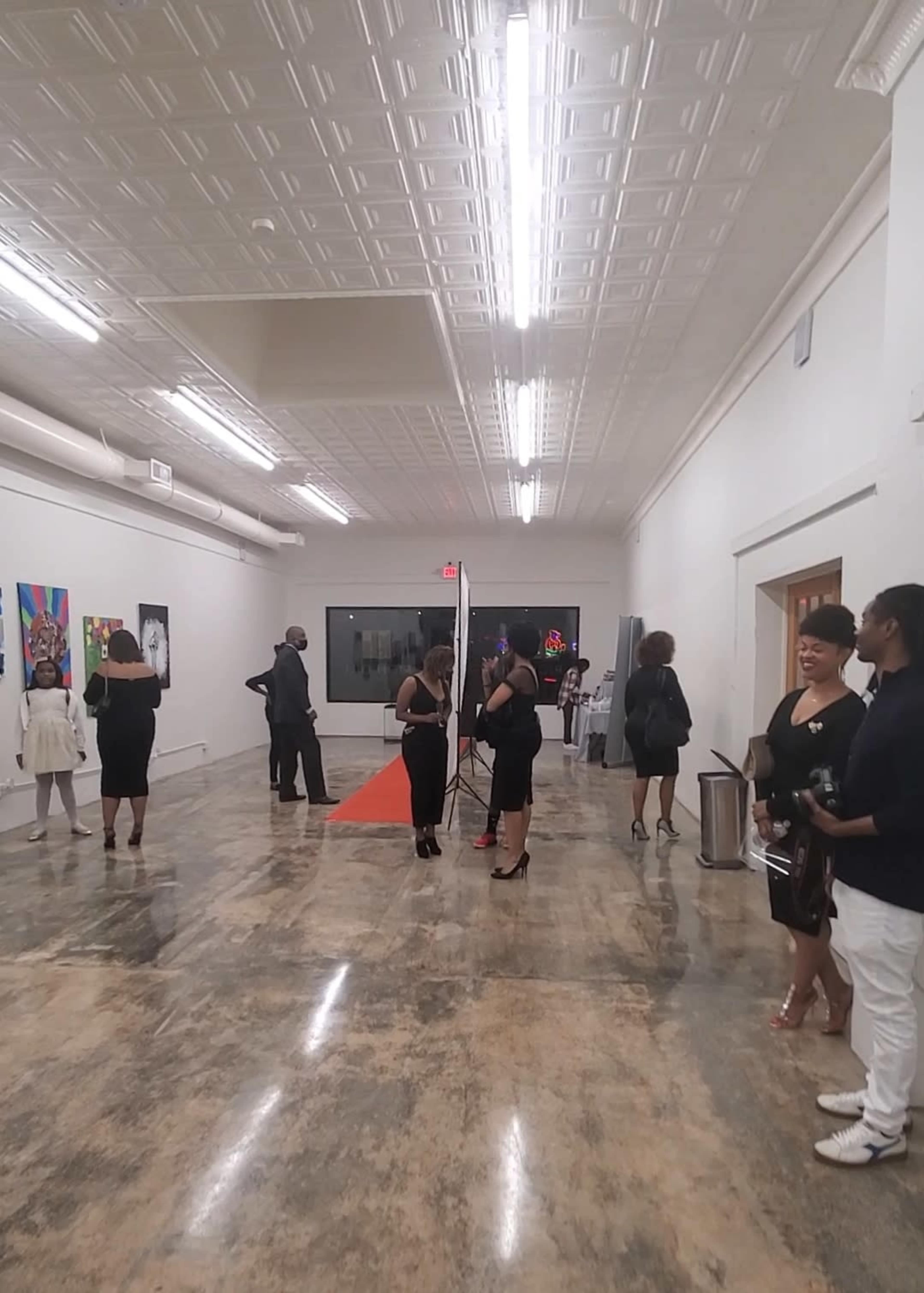 The image shows a group of people in formal attire gathered in a spacious gallery with colorful artwork displayed on the walls.