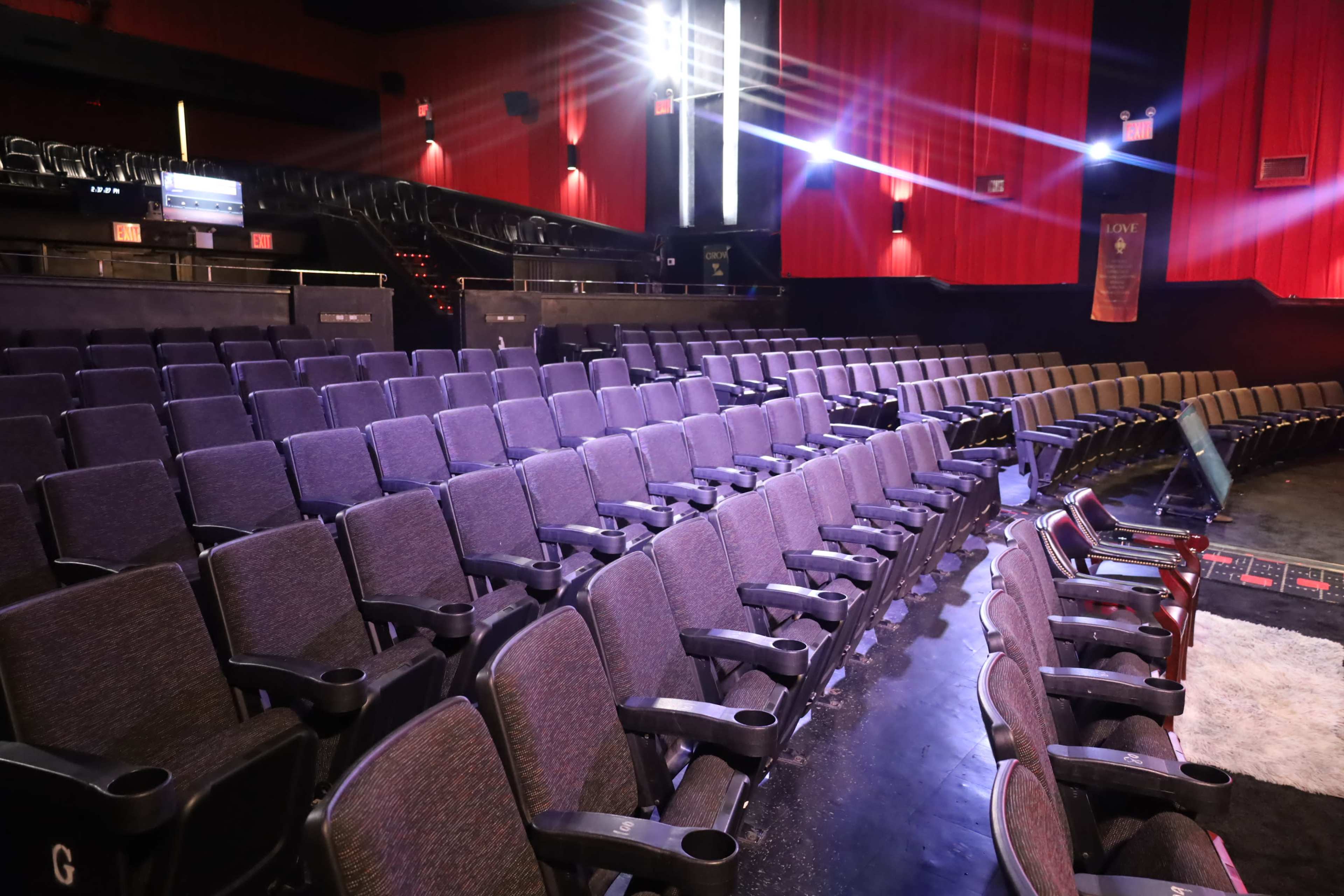 The image shows a dimly lit theater with rows of empty purple seats and a red curtain backdrop.