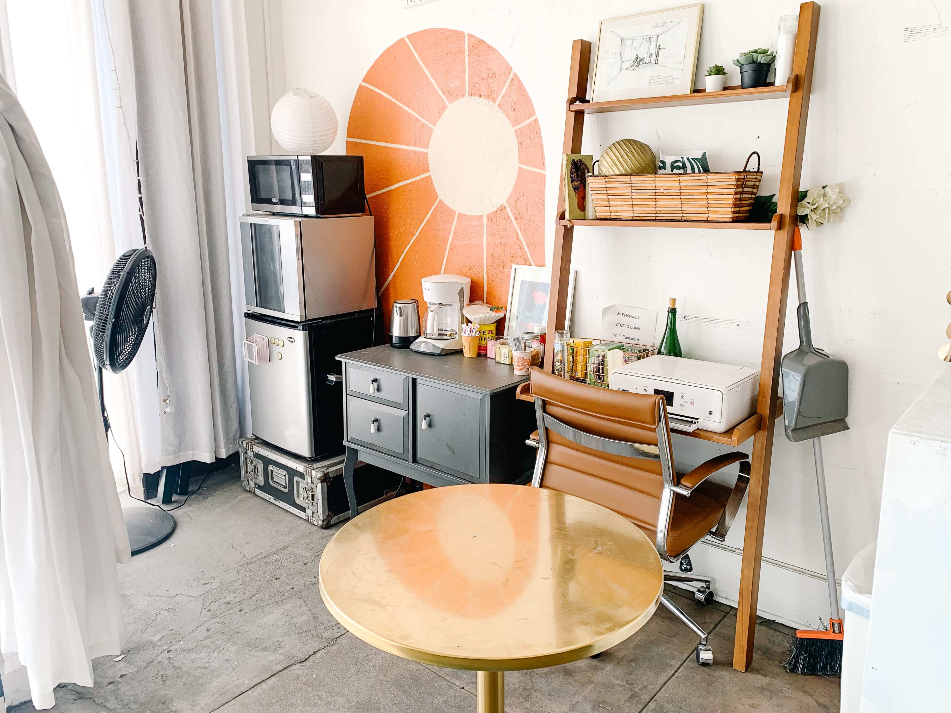 A kitchen corner with a round table, a wooden shelving unit with various kitchen items, a small refrigerator, a coffee maker, and a fan.
