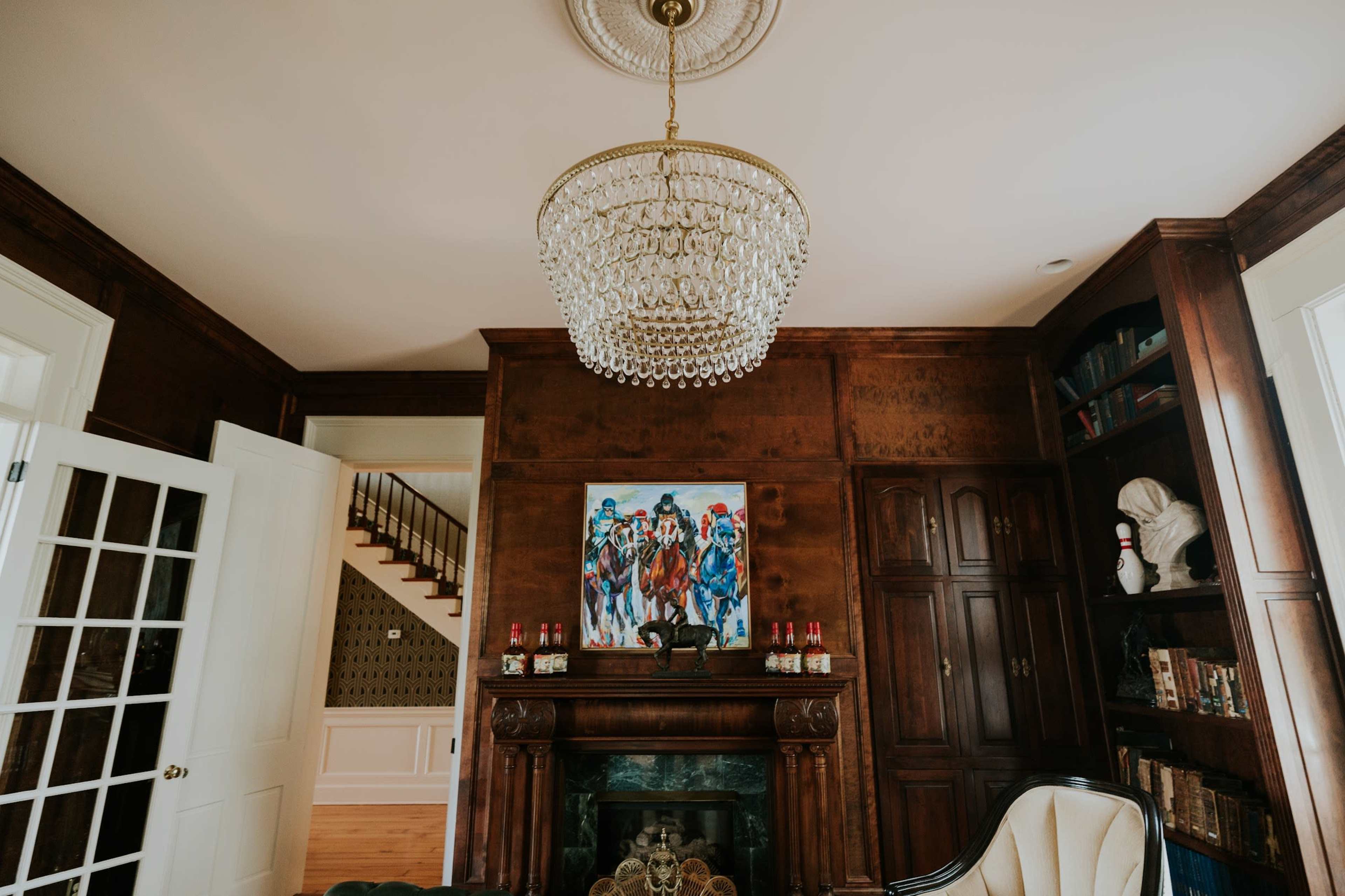 The image shows a warmly lit room featuring a large chandelier, a fireplace adorned with a painting, and wooden shelving filled with books.