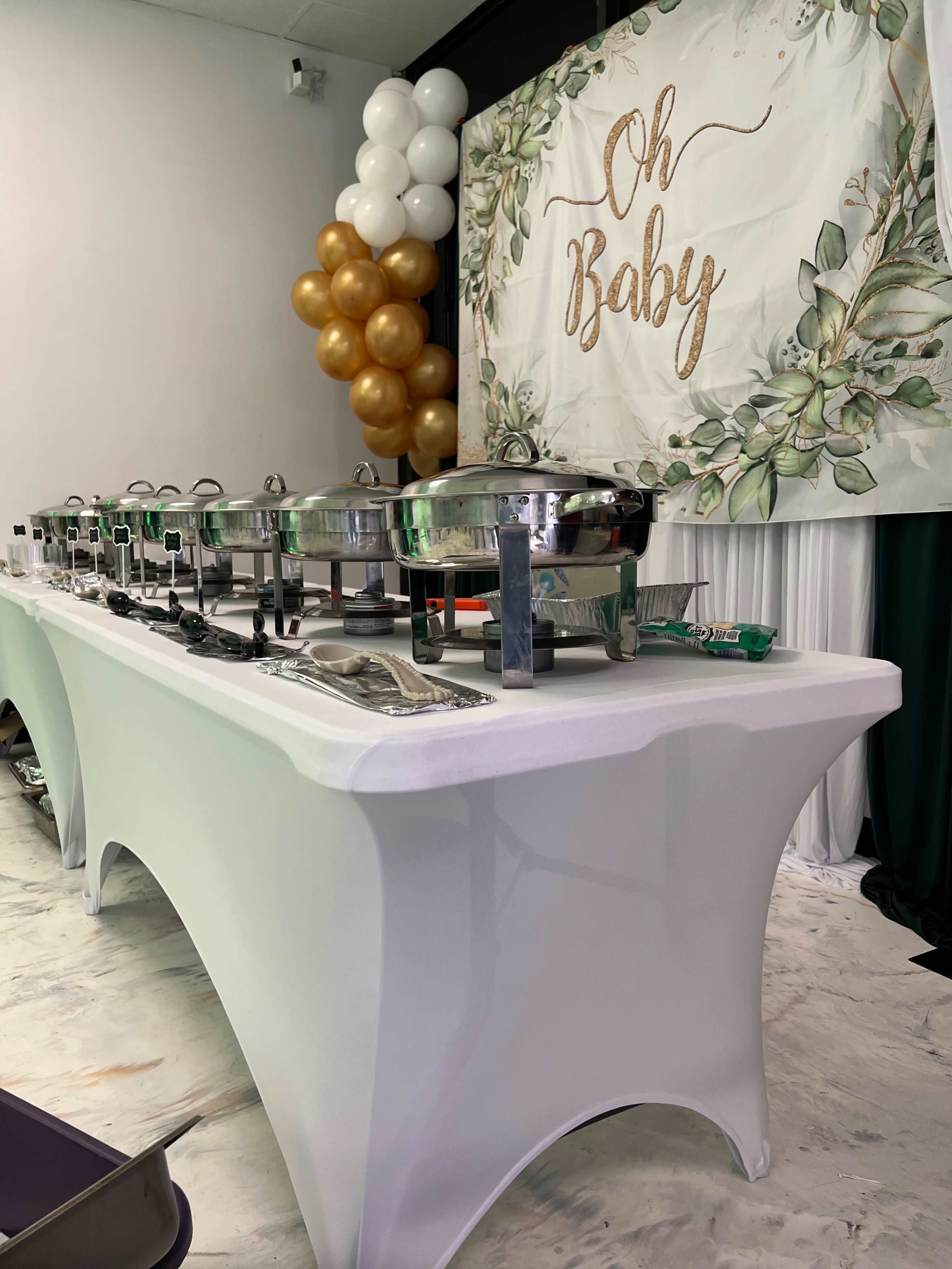 A buffet table is set up with chafing dishes, accompanied by a backdrop featuring the text "Oh Baby" and decorative balloons in gold and white.
