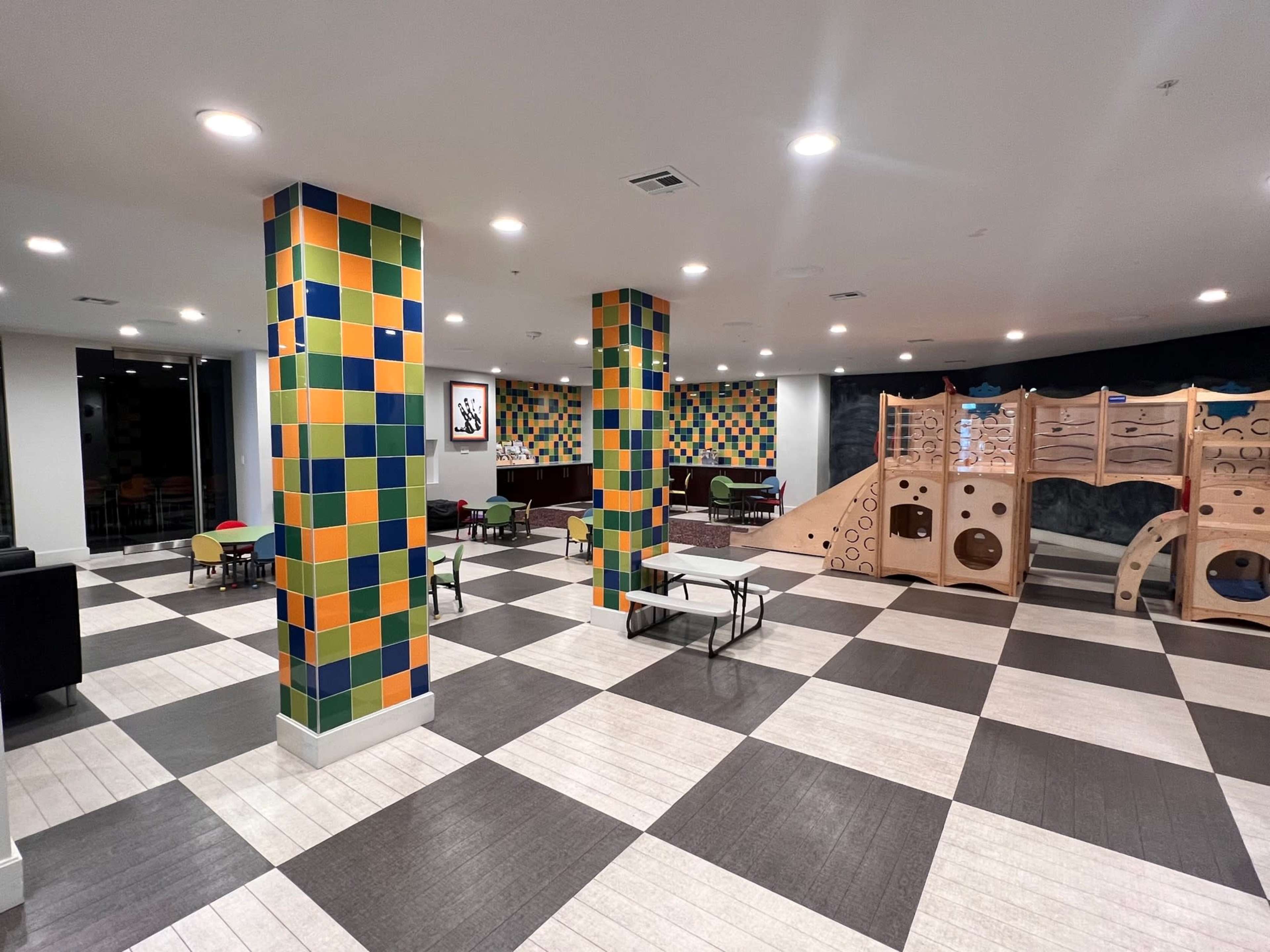 A spacious indoor play area with colorful tiled pillars, a large play structure, and seating arranged in a checkerboard floor pattern.