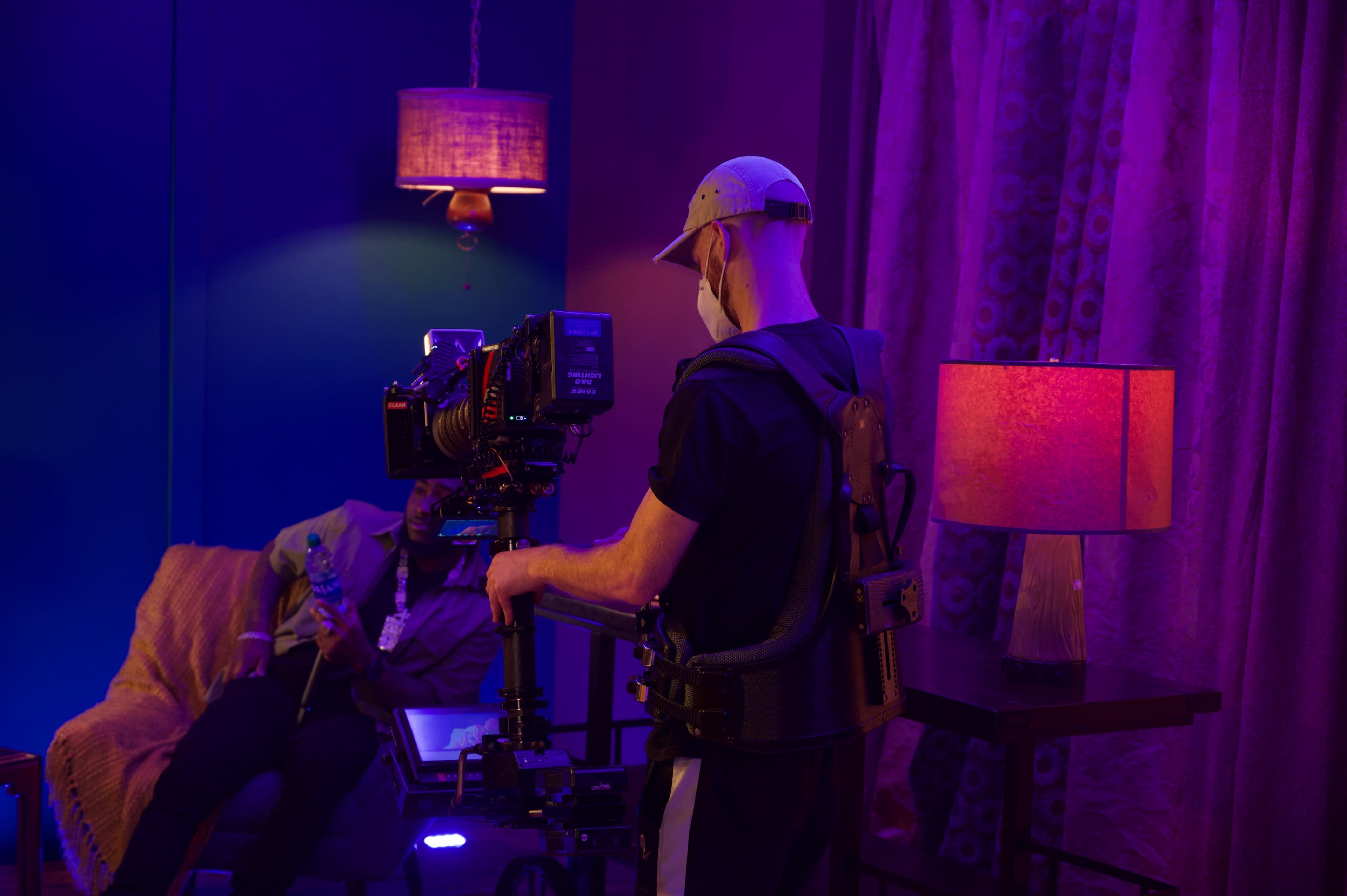 A camera operator uses a rig to film a scene featuring a seated subject in a dimly lit room with colorful lighting.