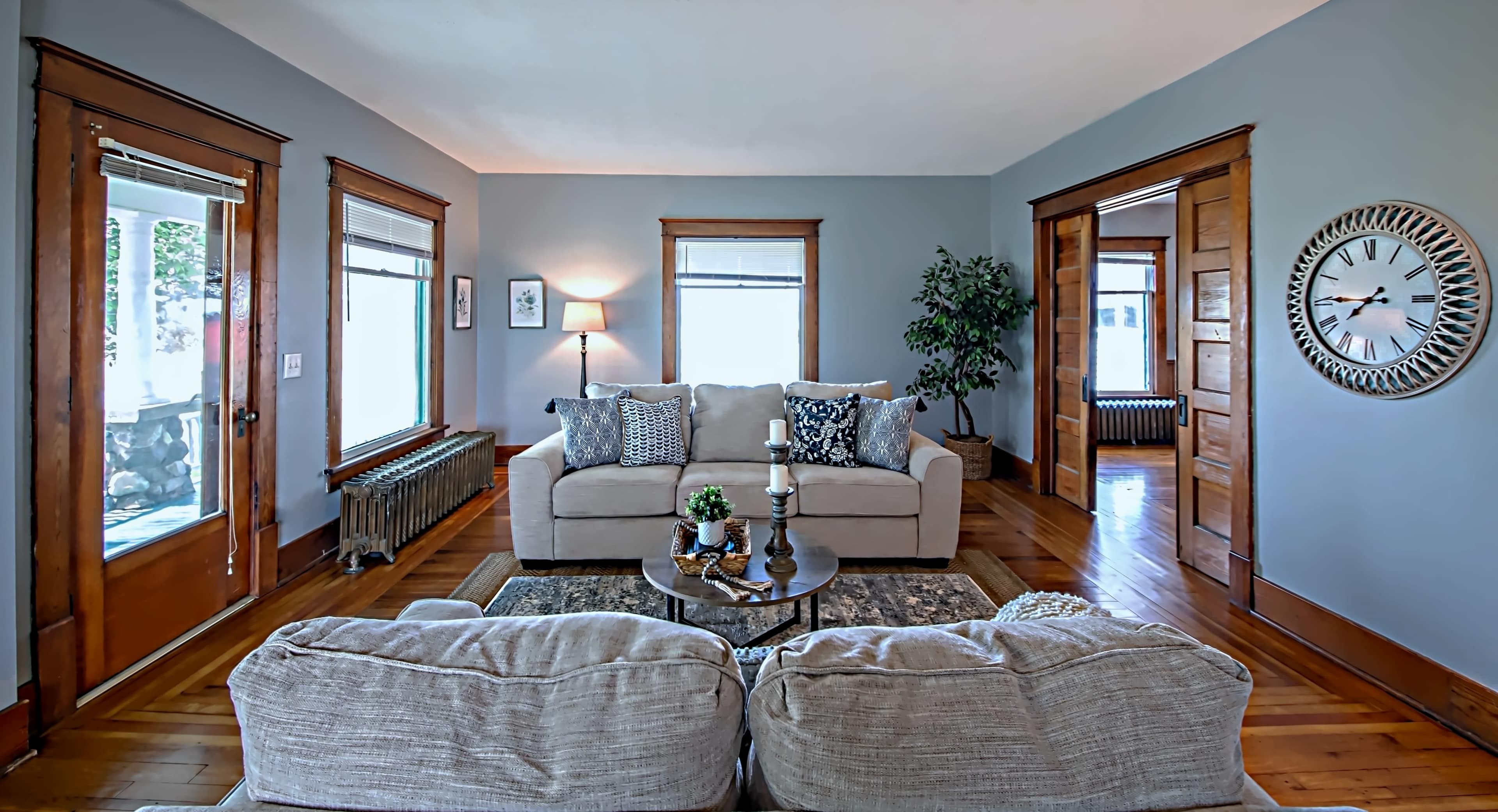 A living room features a light-colored sofa with patterned cushions, surrounded by hardwood floors and large windows, accented by wooden door frames and a wall clock.