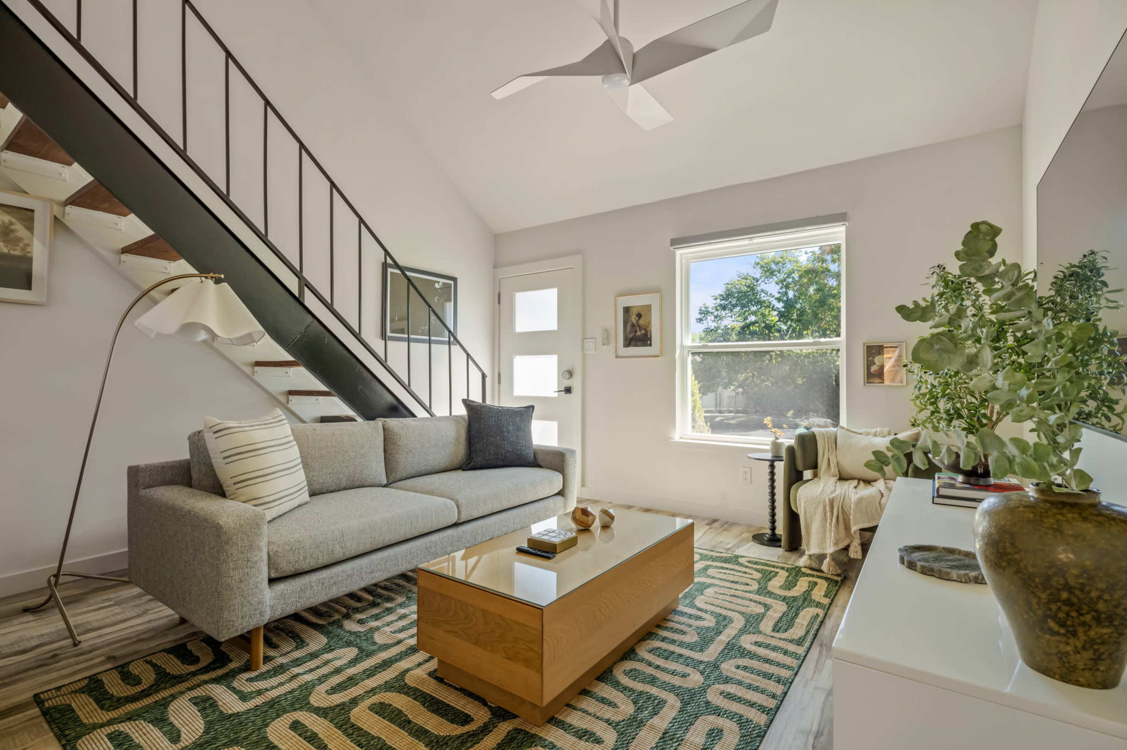 The image depicts a modern living room with a gray sofa, a wooden coffee table, a patterned rug, and a staircase in the background.
