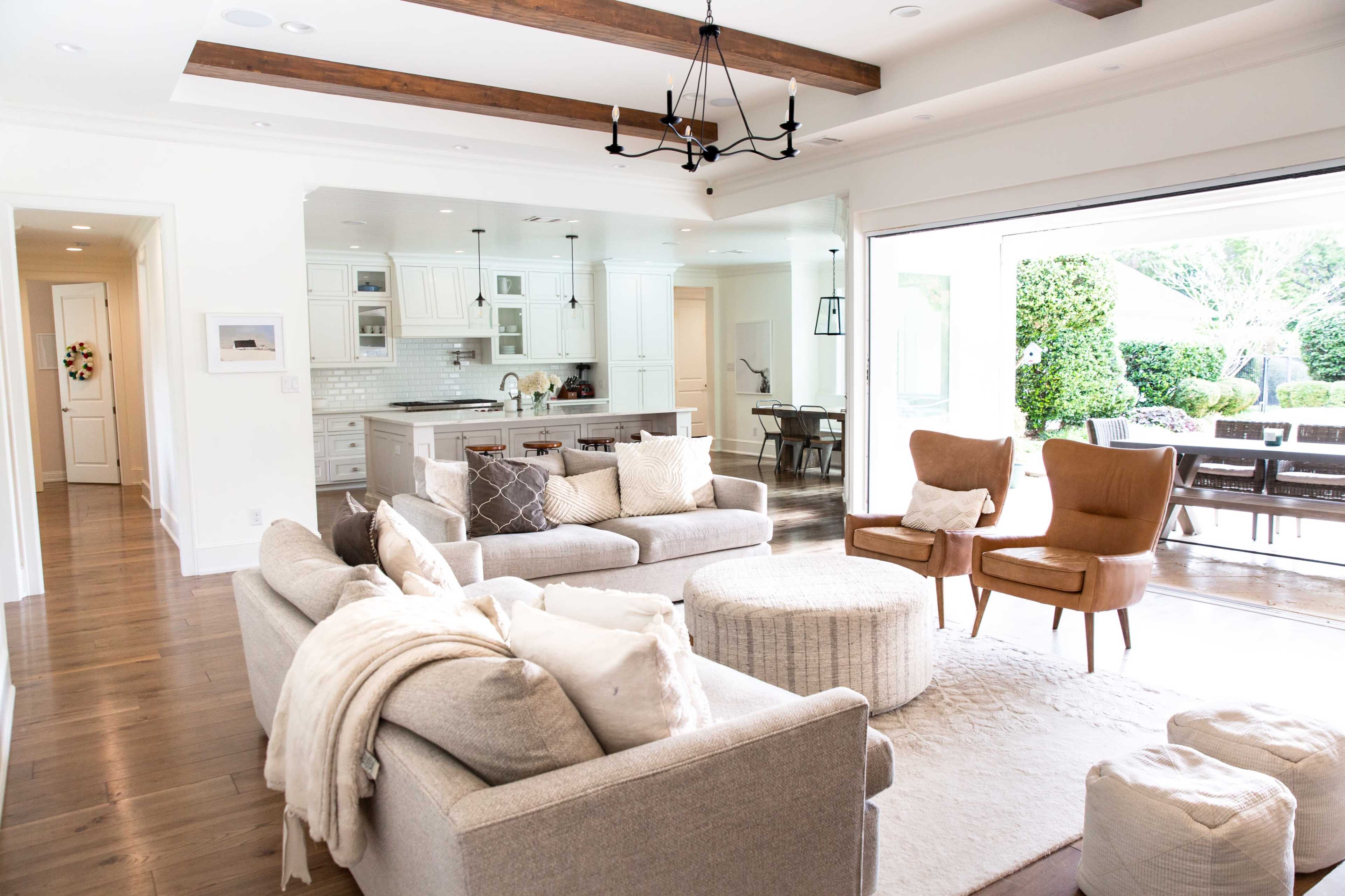 A bright, modern living room with a light-colored sofa, two brown armchairs, and a round ottoman, leading into a kitchen and dining area through an open layout.