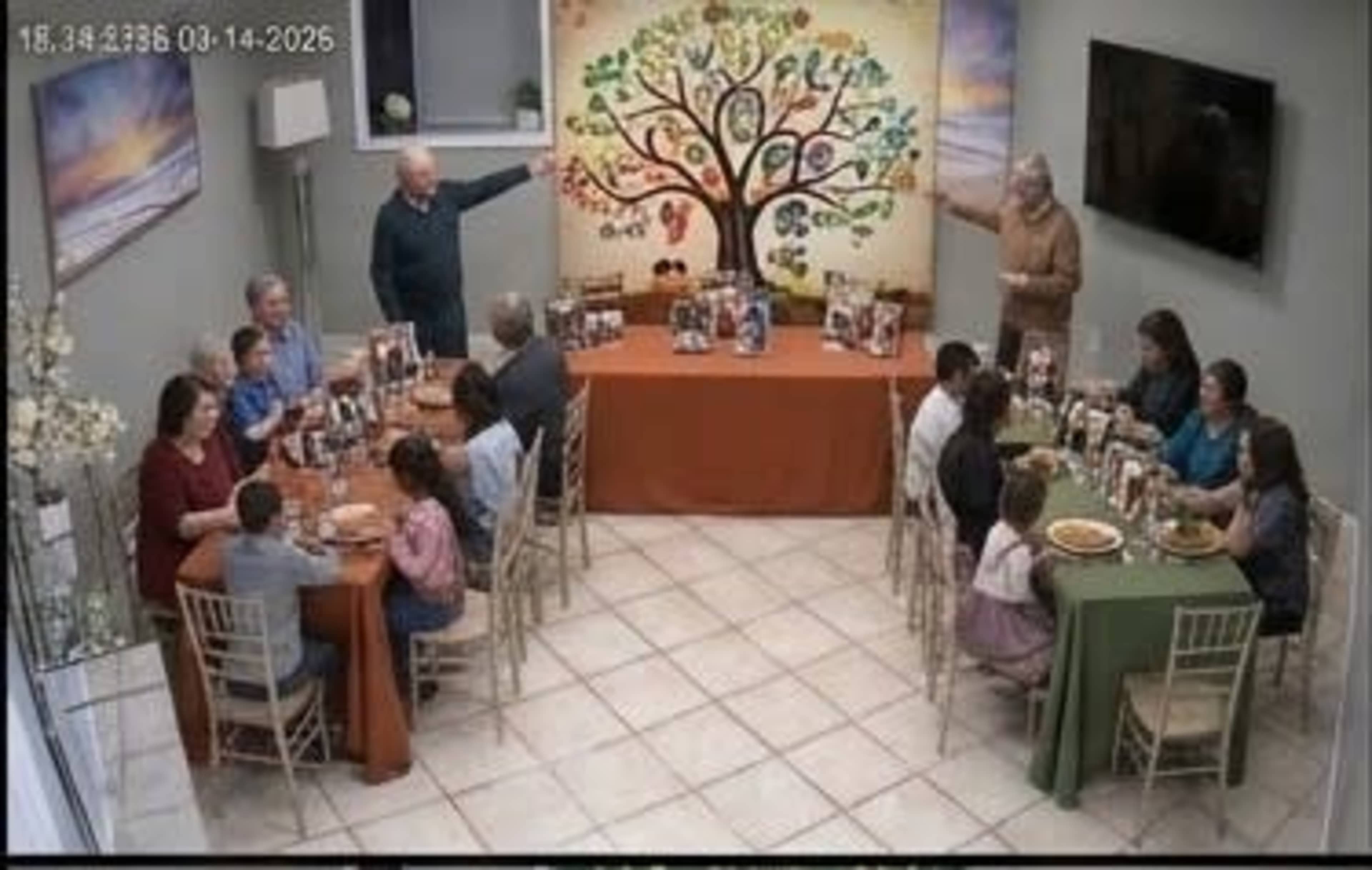 A large gathering of people sits around tables in a decorated room, with a colorful tree mural on the wall and various dishes displayed on the tables.