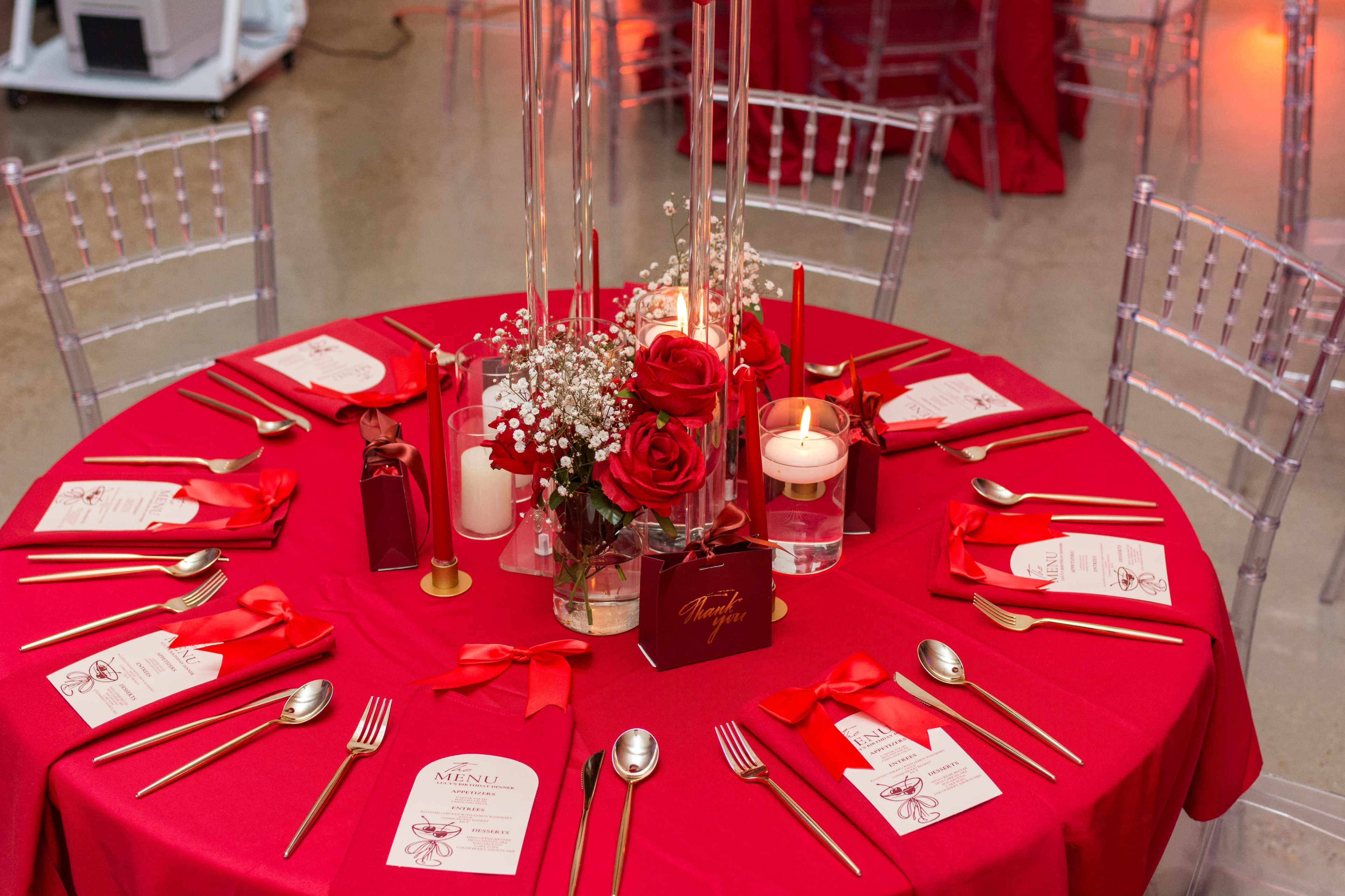 The table is set with a red cloth, golden cutlery, and decorative centerpieces featuring roses, candles, and elegant place cards.